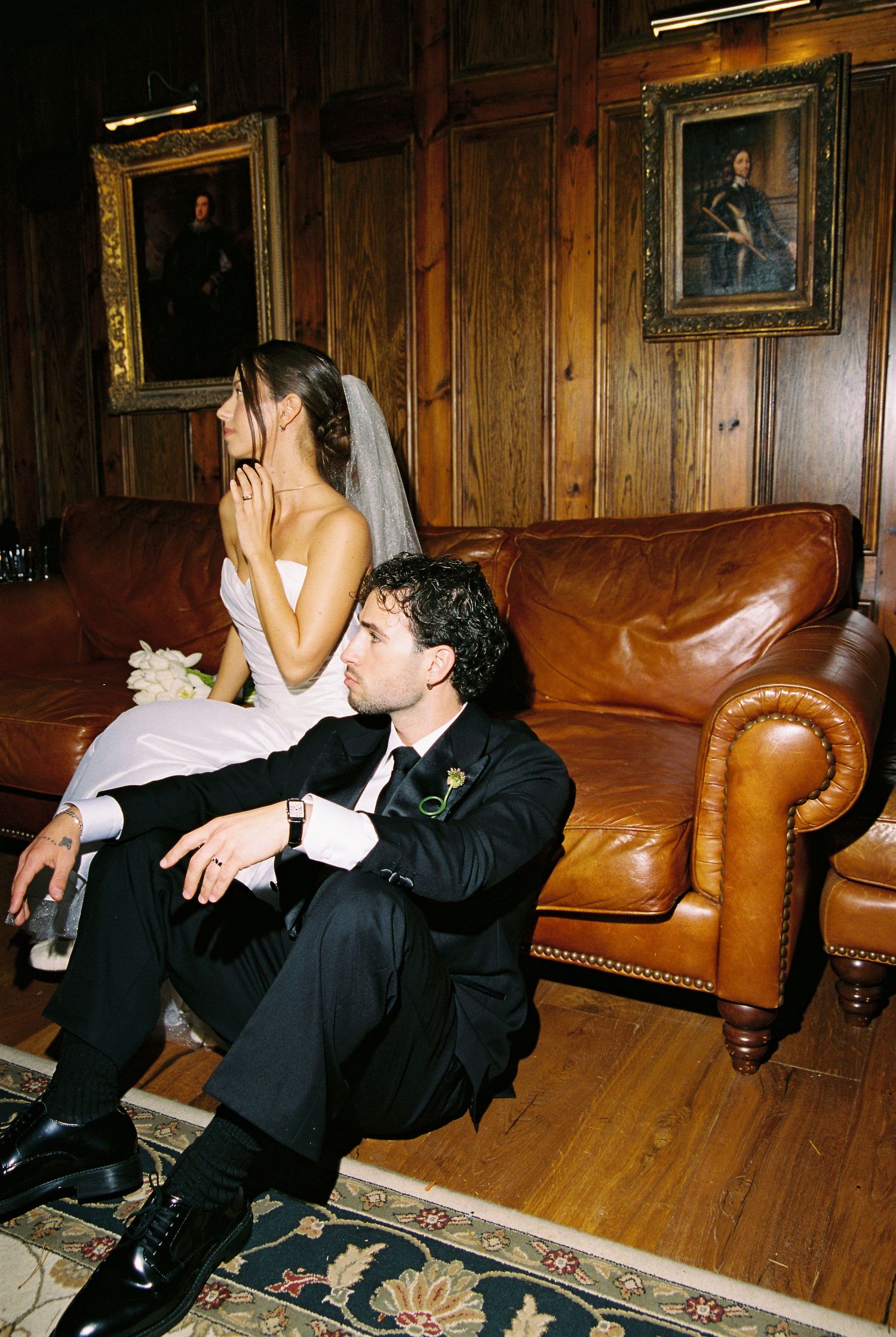 Bride in white gown and groom in tuxedo sitting apart on leather sofa in wood-paneled room.