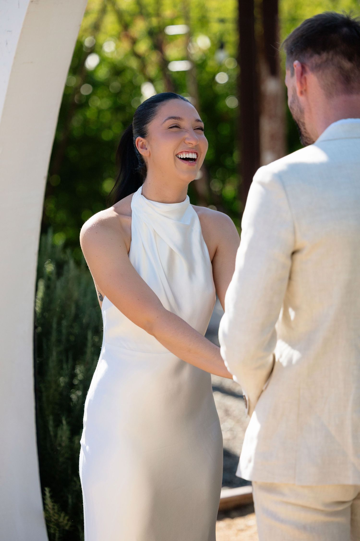 Joyful bride in white satin halter dress laughs while holding groom's hands at outdoor wedding ceremony.