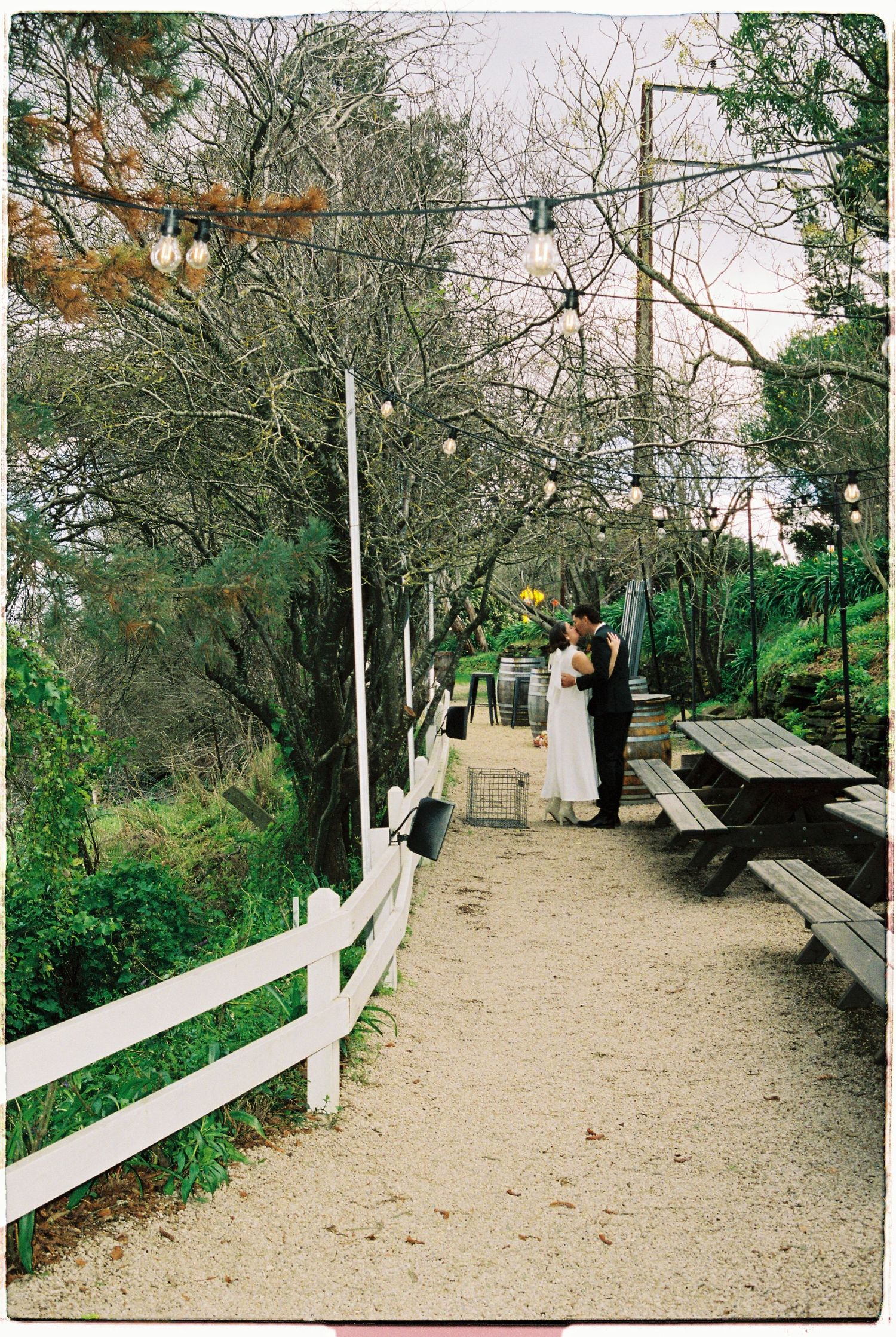 A bride and groom share a romantic moment on a garden path lined with string lights and picnic tables.