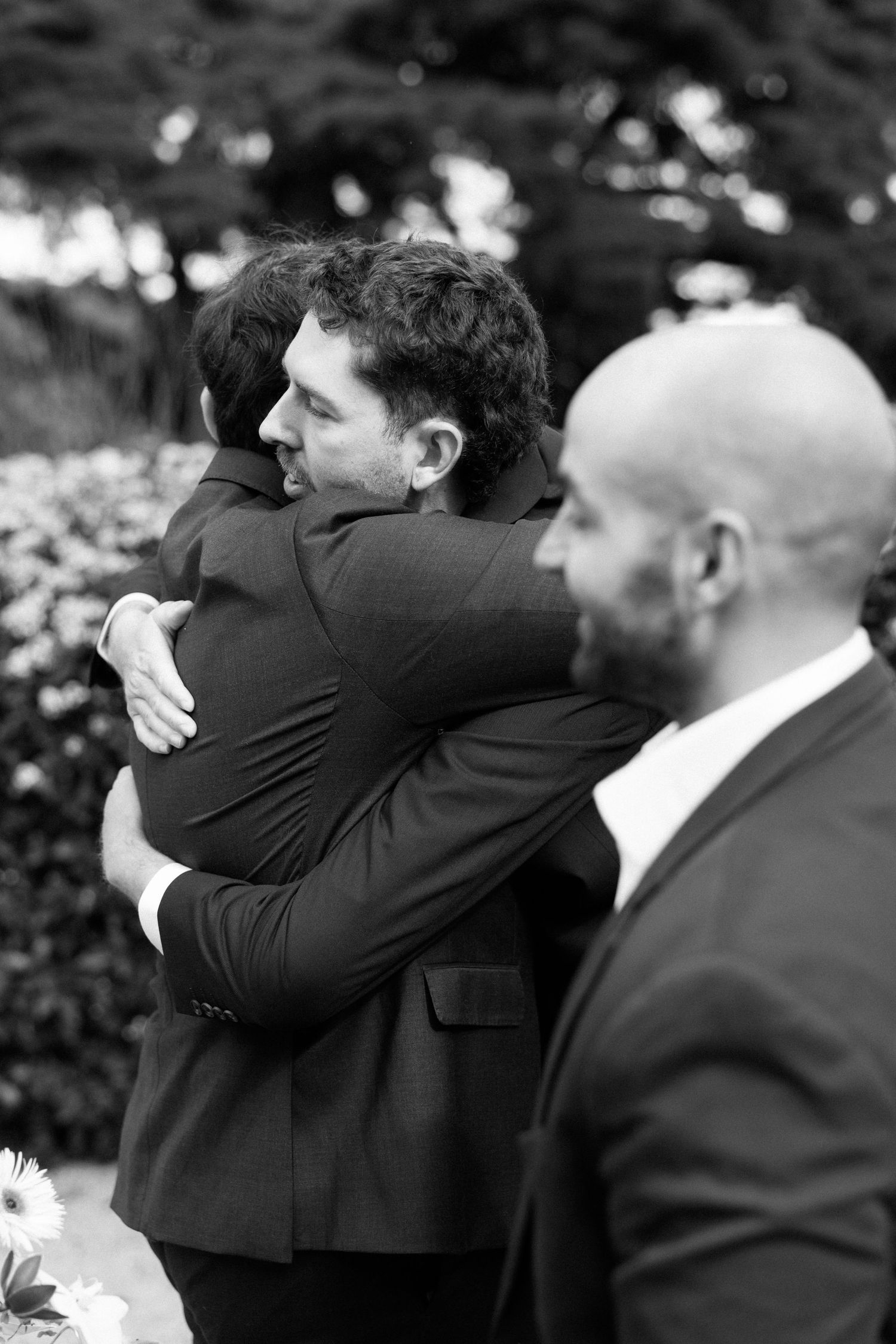 Two men in suits share an emotional hug at a wedding ceremony, with a bald bearded man standing nearby.