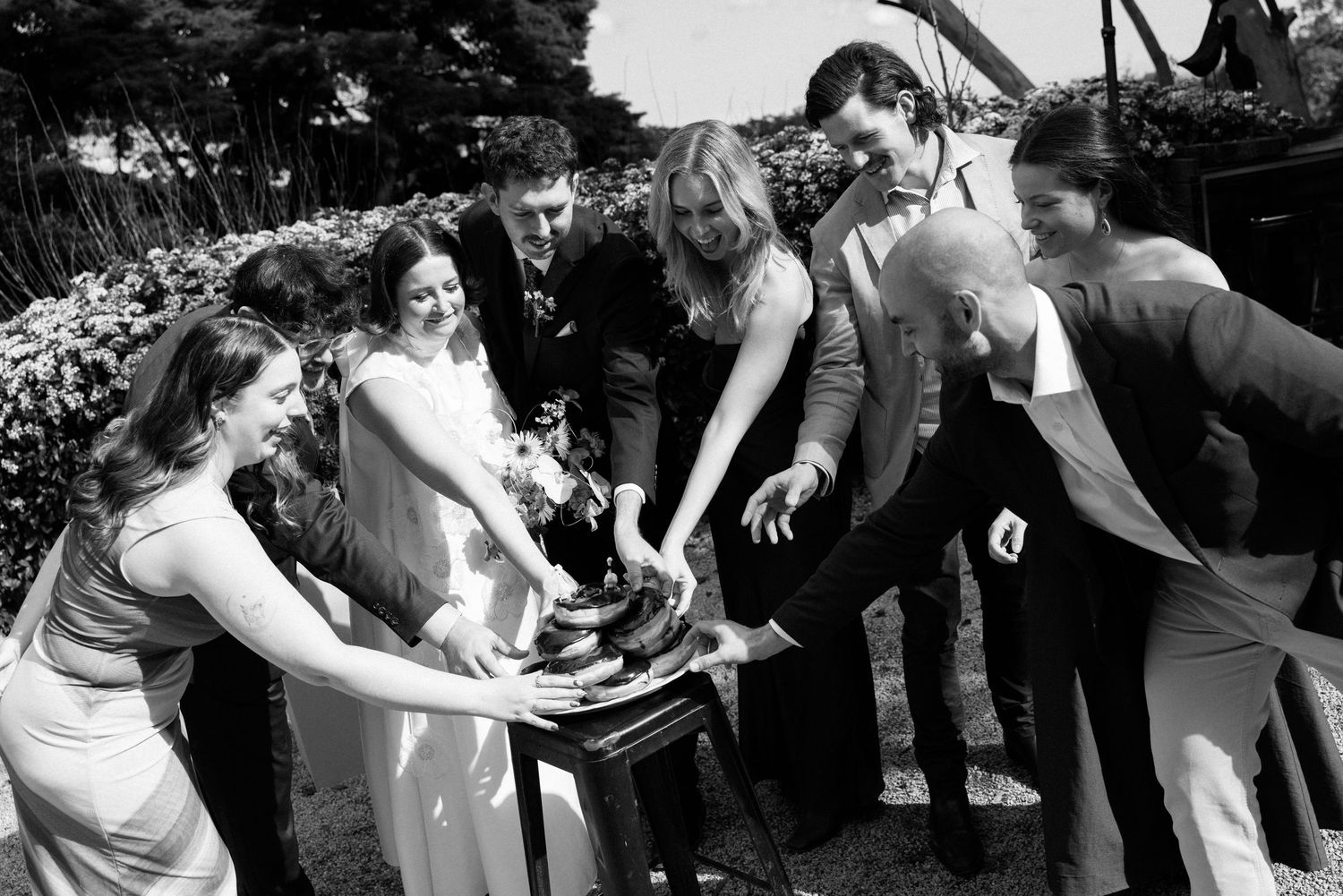 Black and white wedding photo of bride, groom, and guests gathered around a table cutting cake outdoors.