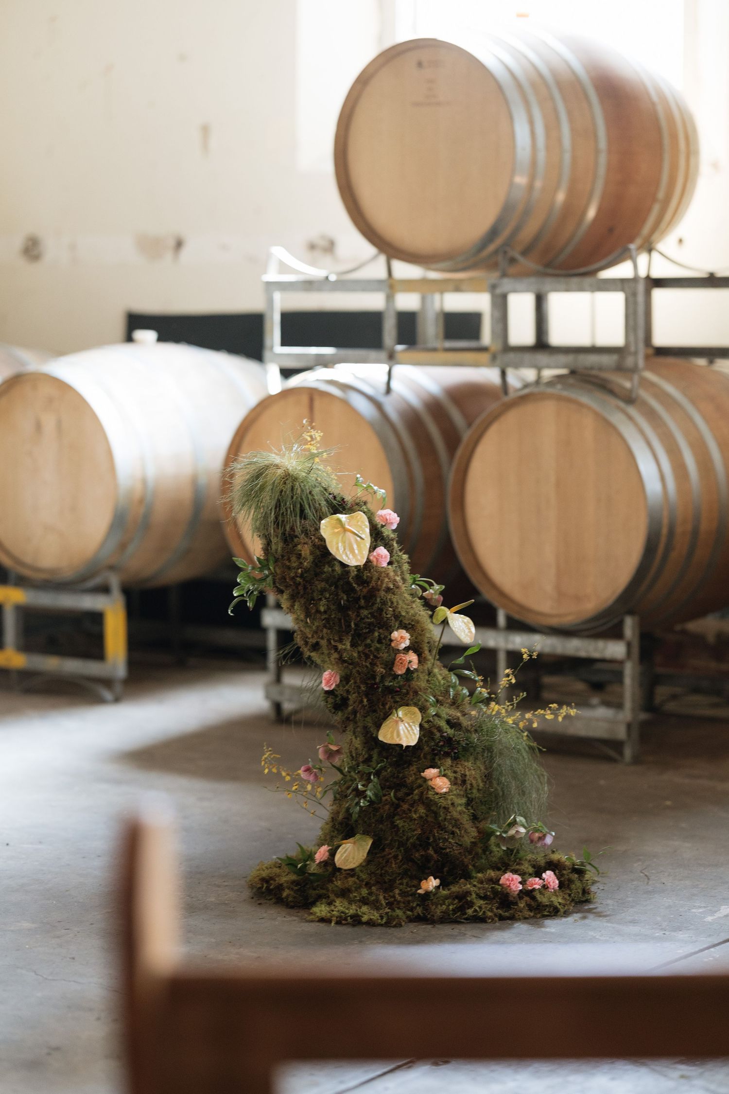 Moss floral arrangement with pink roses and anthuriums displayed among oak wine barrels in a winery.