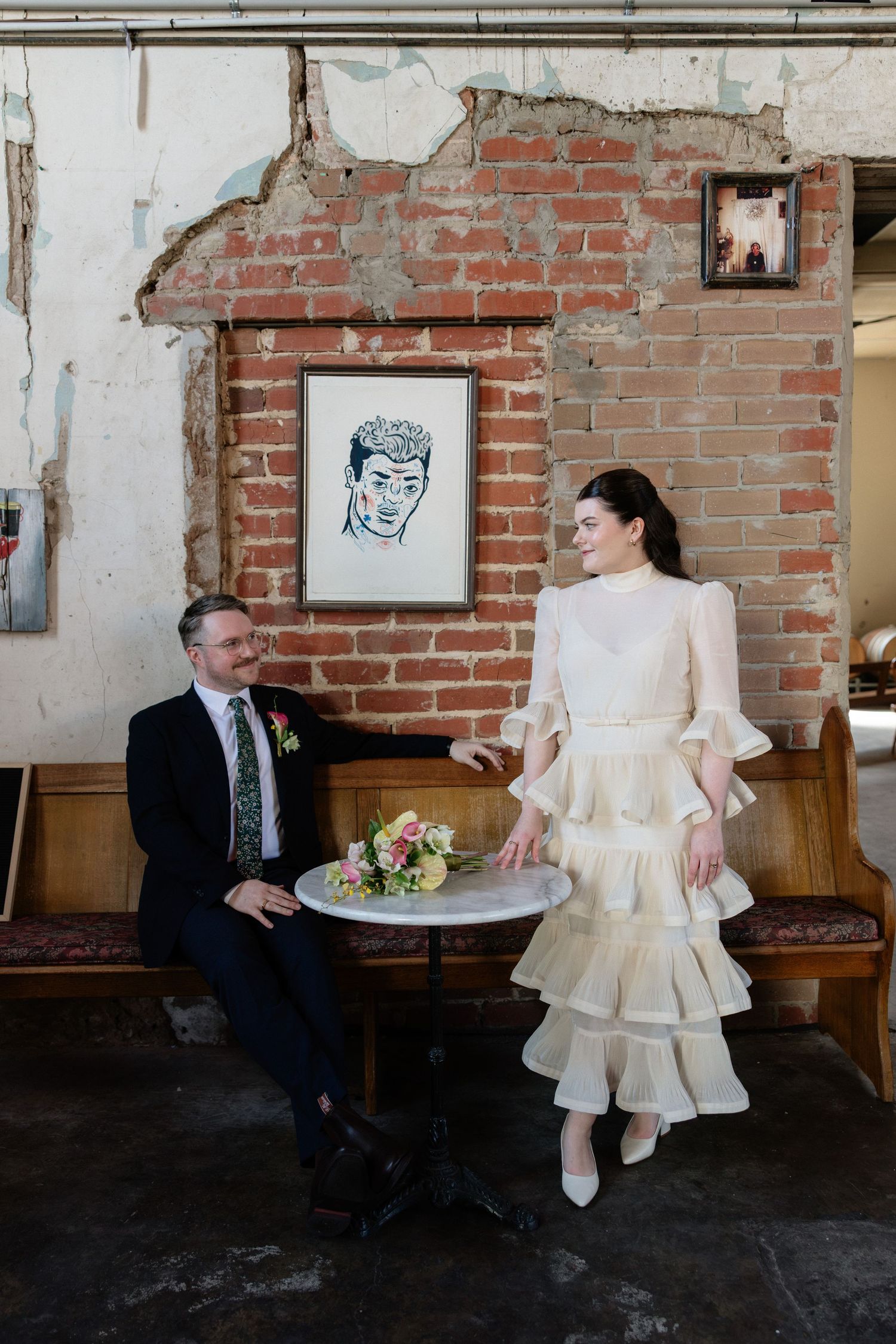 Bride in tiered white dress and groom in navy suit share a joyful moment by a brick wall with framed artwork.