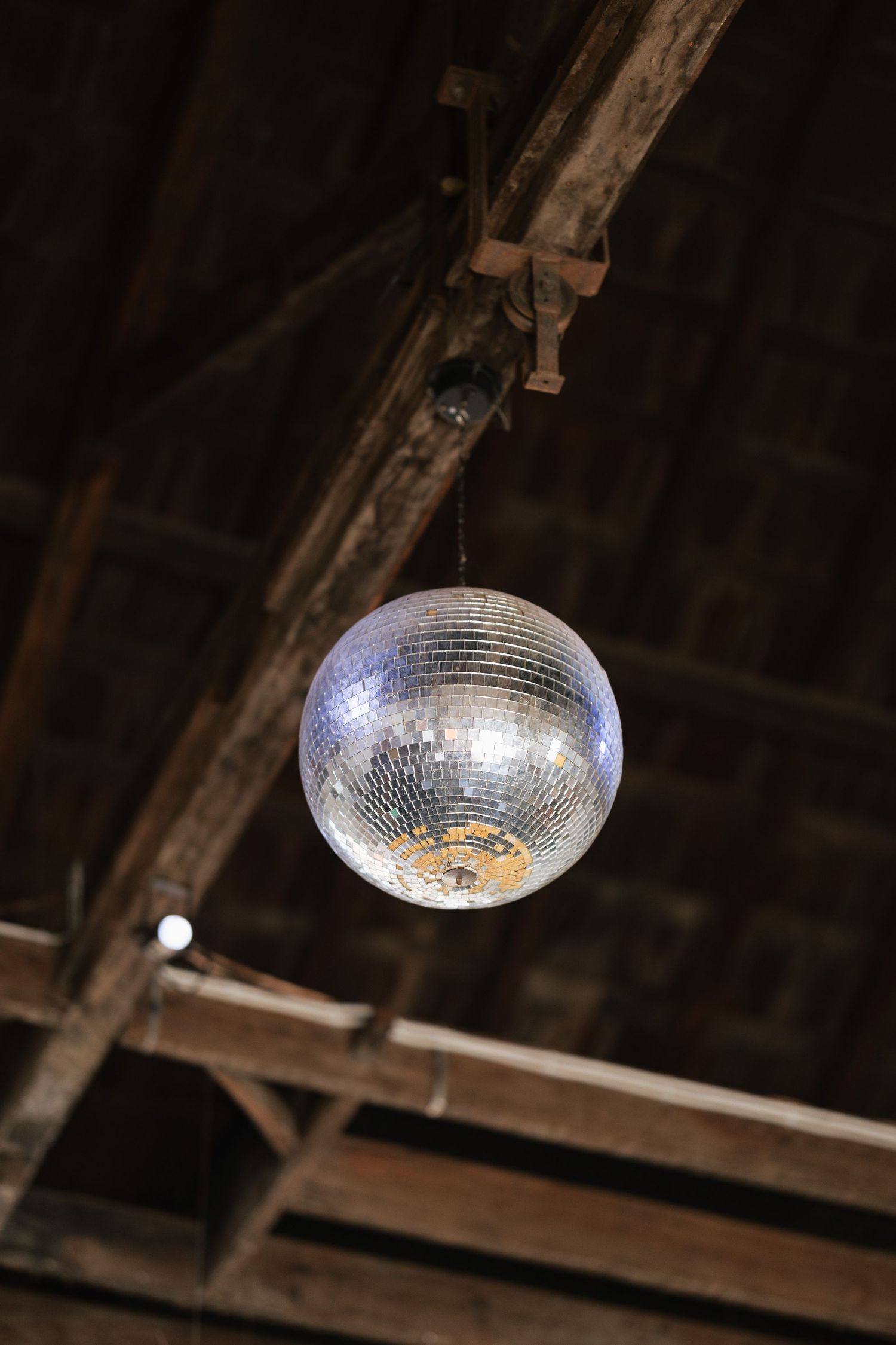 Shimmering disco mirror ball hanging from rustic wooden beam ceiling in a dimly lit venue.