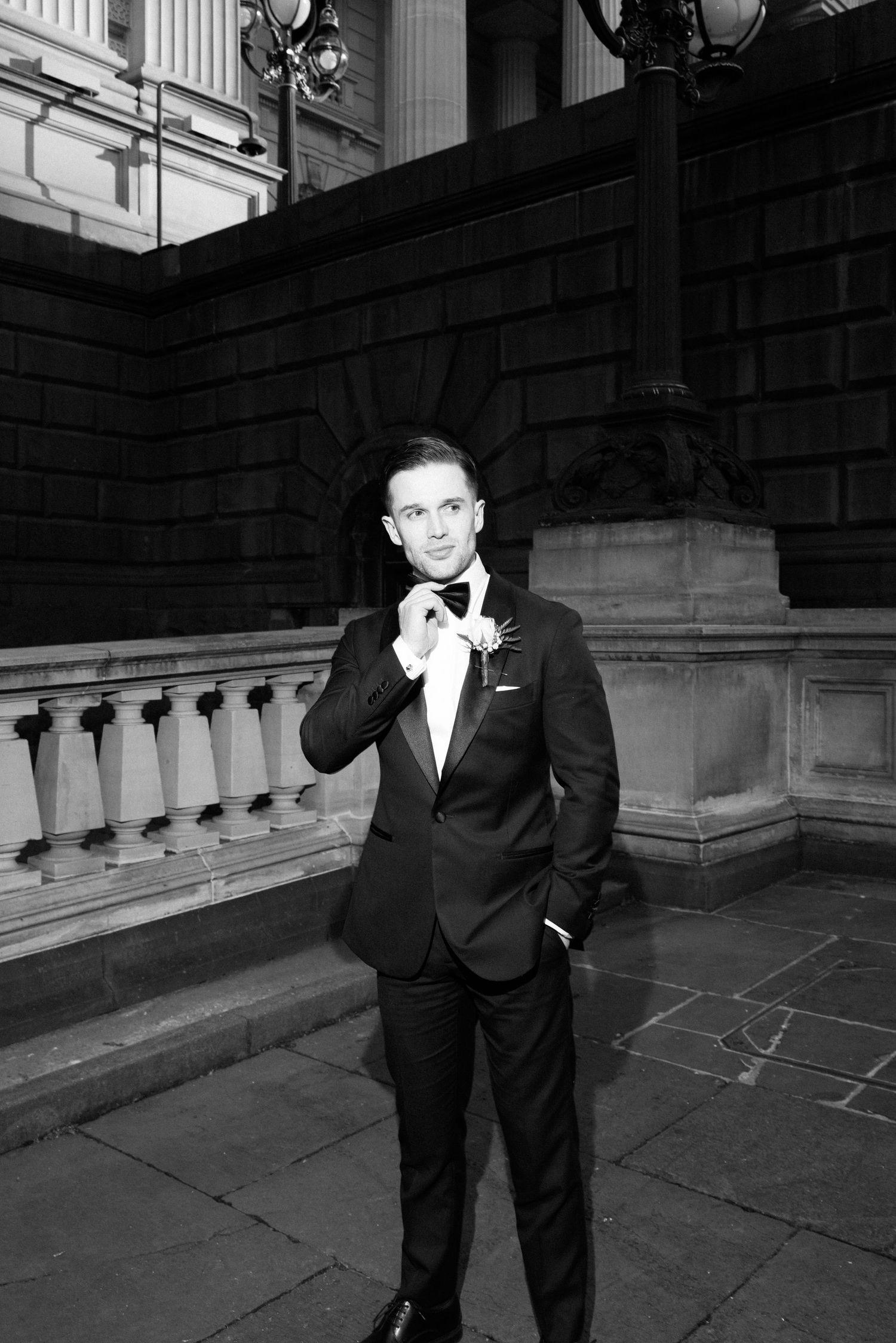 Dapper groom in black tuxedo adjusting bow tie outside grand neoclassical building in black and white.
