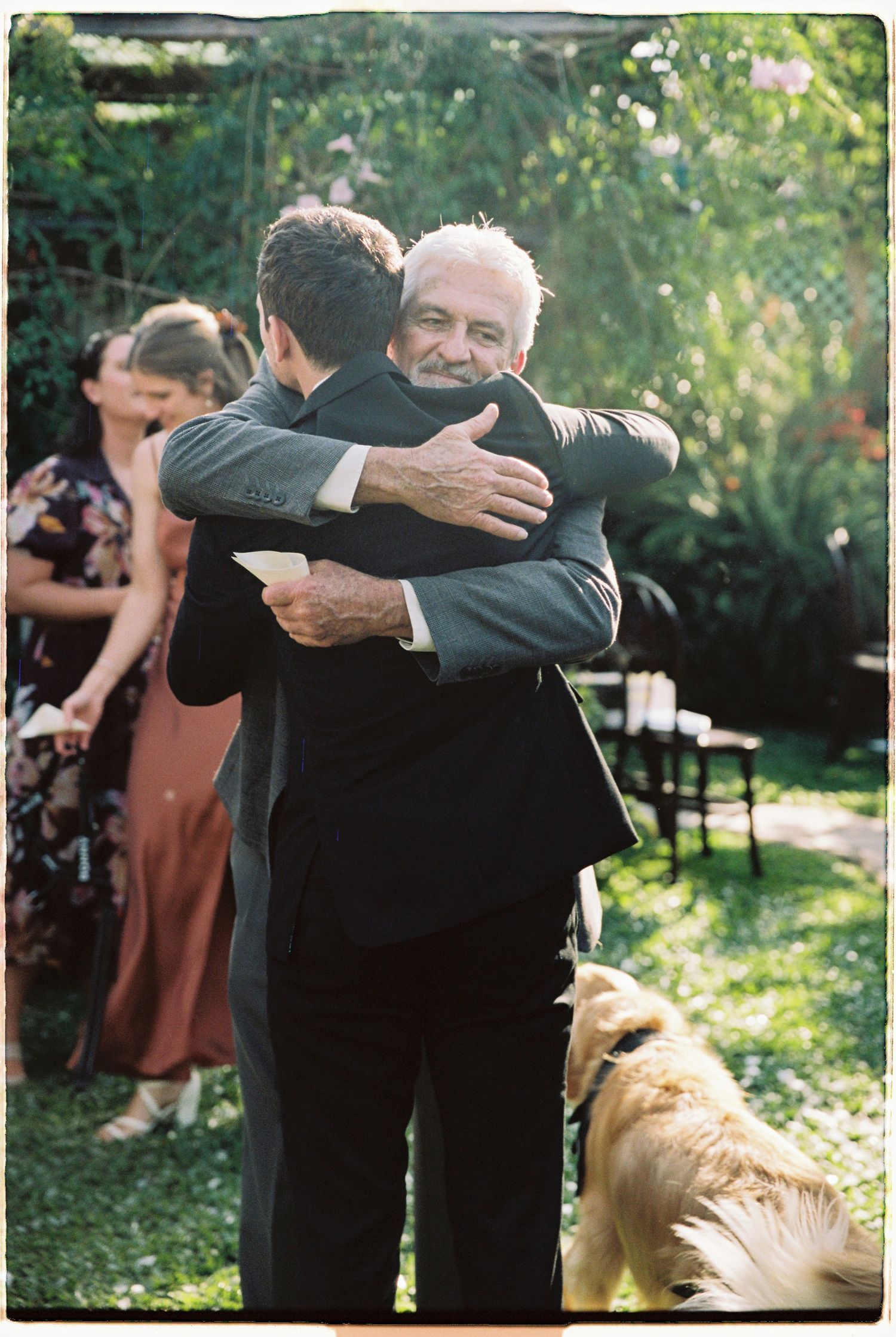 Two men in suits share a warm hug at an outdoor wedding reception with guests and a golden retriever nearby.