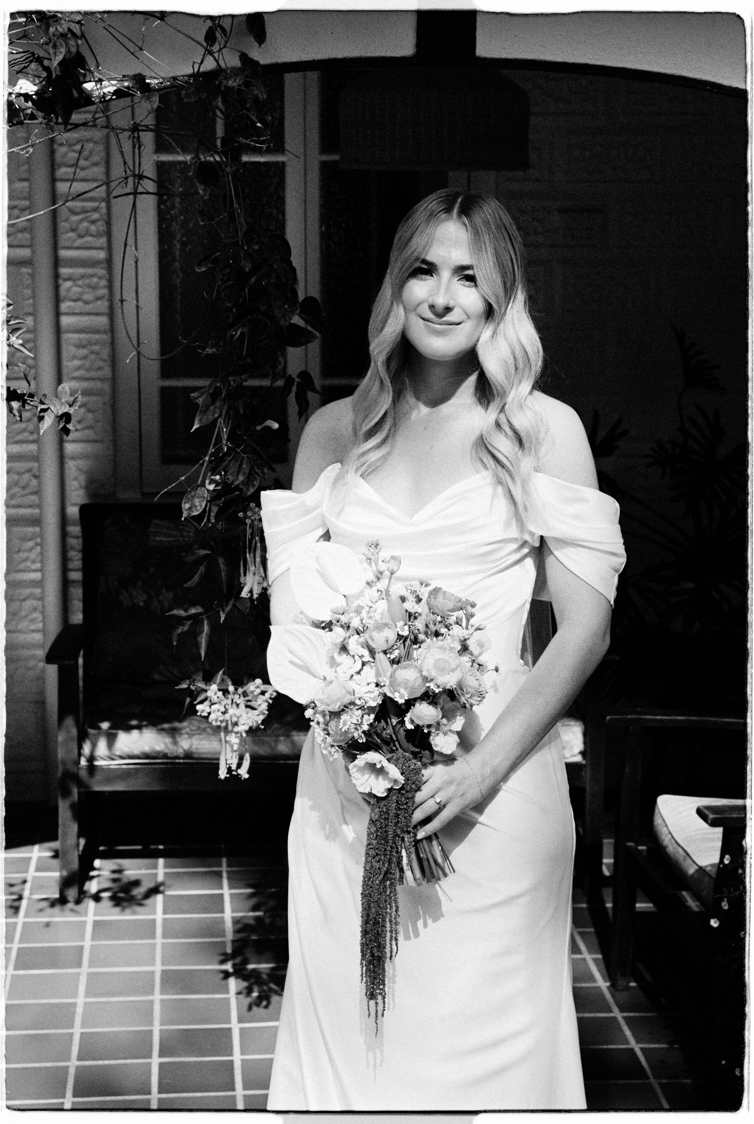 Black and white portrait of a smiling bride in off-shoulder gown holding a floral bouquet.