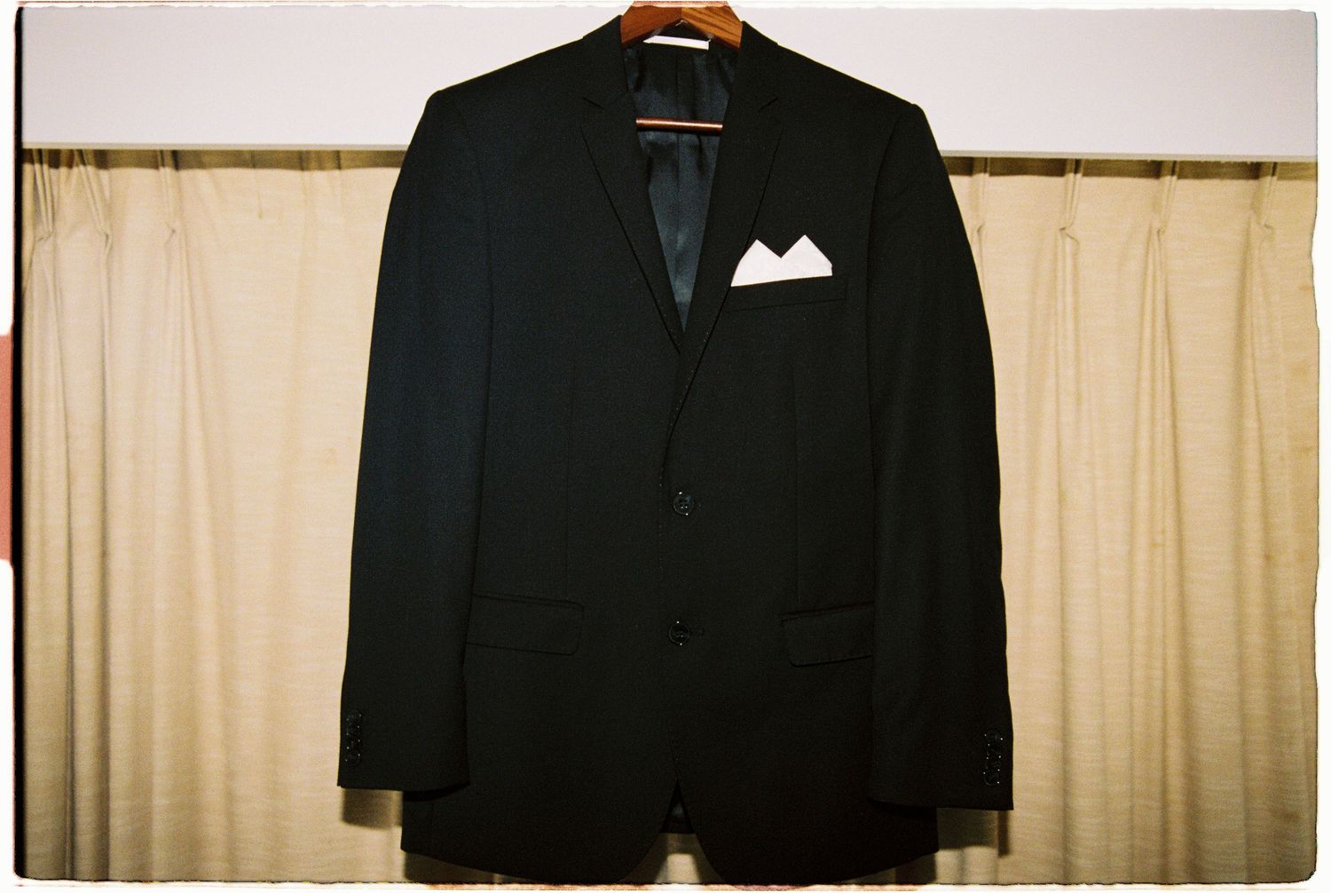 Black suit jacket with white pocket square on wooden hanger against beige curtain backdrop.