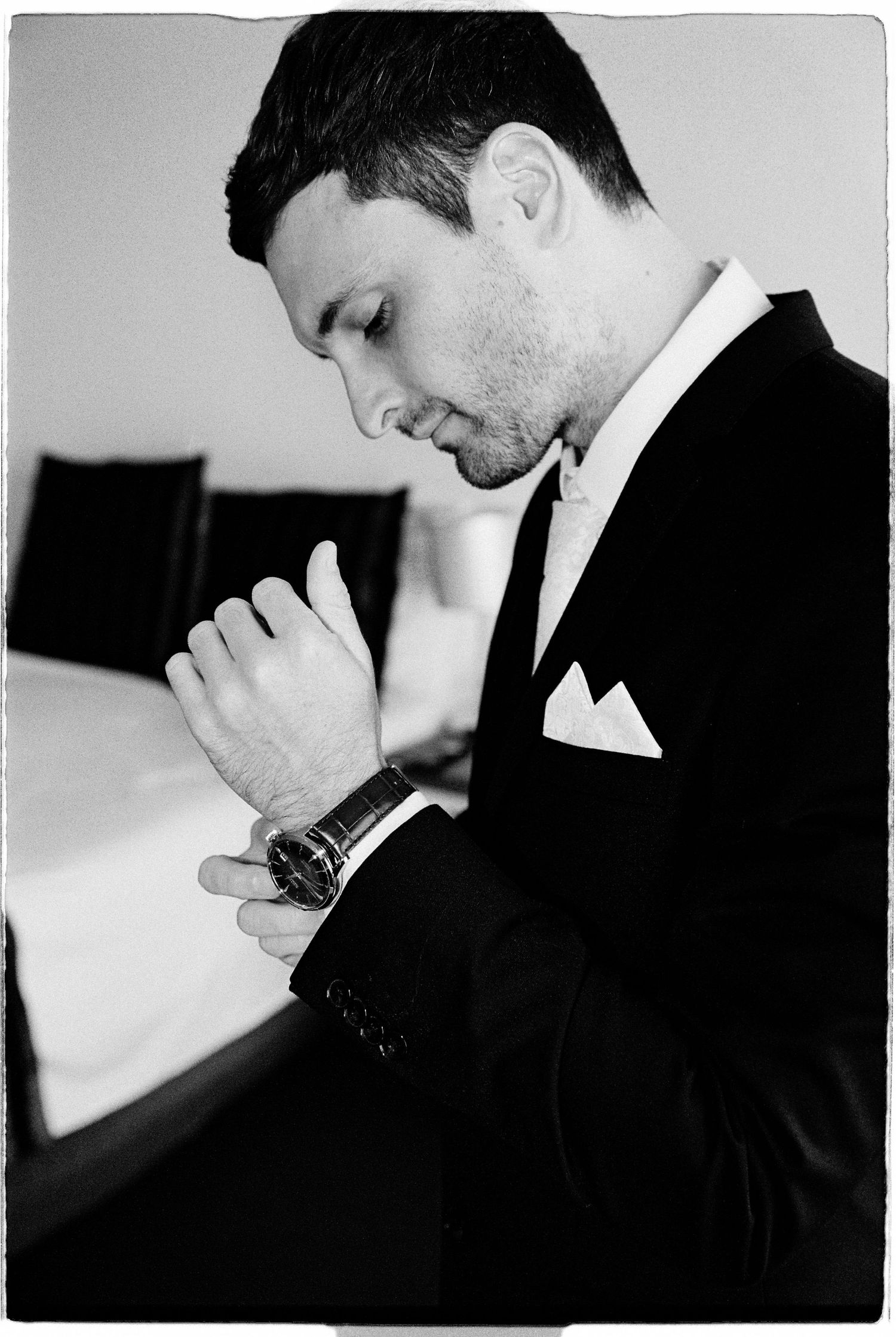 Black and white photo of an elegant man in a tuxedo adjusting his watch, with a pocket square visible.