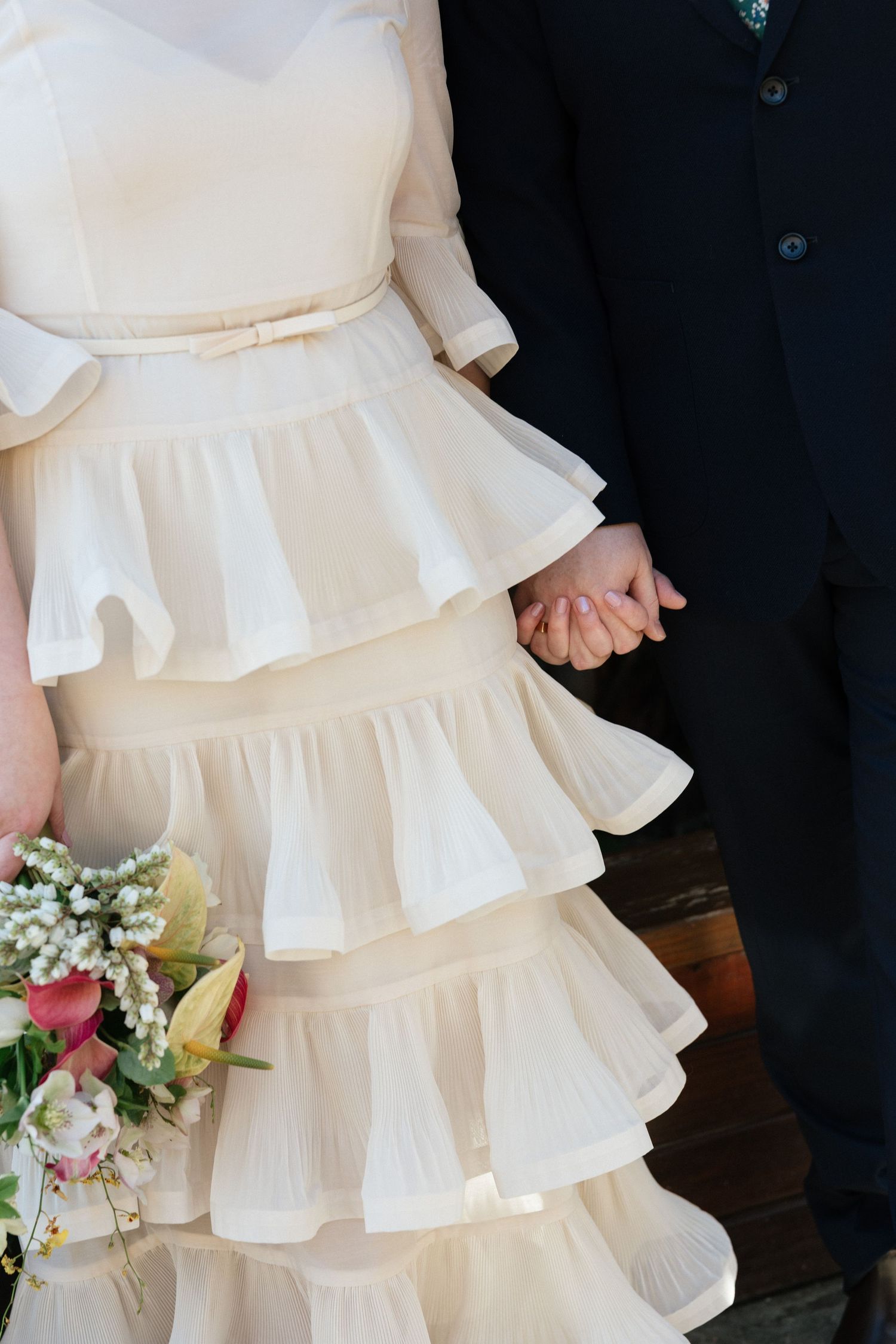 Bride in tiered ruffle wedding dress holding hands with groom in navy suit, with bridal bouquet of pink flowers.