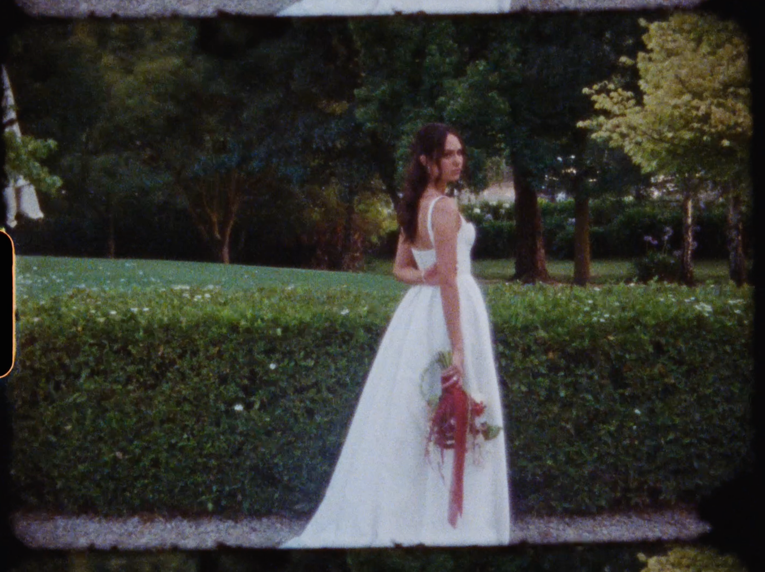Bride in white gown holding colorful bouquet stands in lush green garden, posing elegantly for wedding photo.