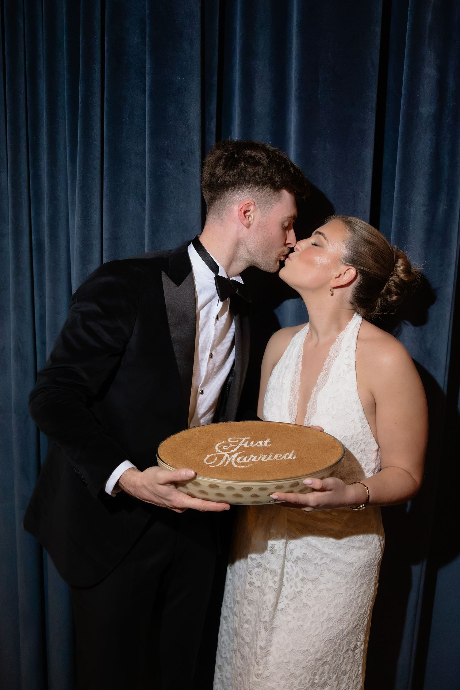 Newlywed couple kissing while holding a wooden Just Married sign, groom in tuxedo and bride in white halter gown.