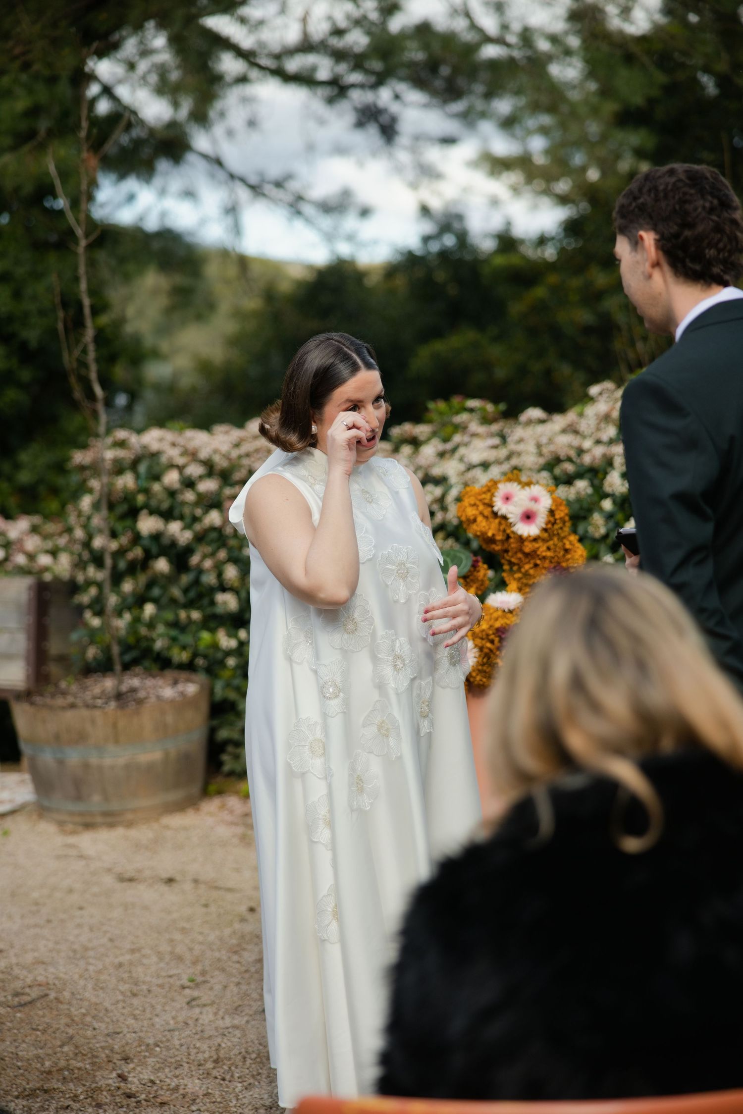 Emotional bride in white lace gown wiping tears during outdoor wedding ceremony surrounded by blooming flowers.