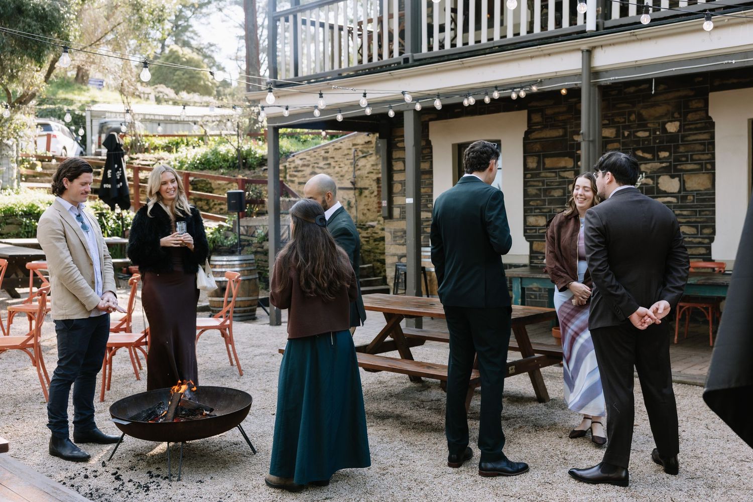 Guests mingle outdoors near a stone building with string lights, a fire pit, and wooden furniture at a casual gathering.