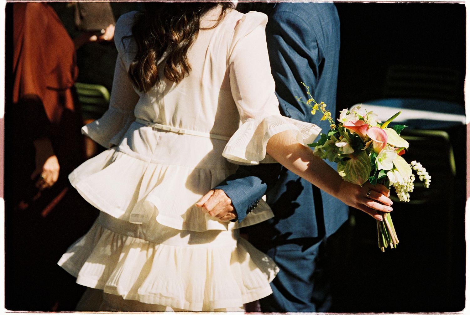 Bride in white ruffled dress holding colorful bouquet, standing beside groom in blue suit at wedding ceremony.
