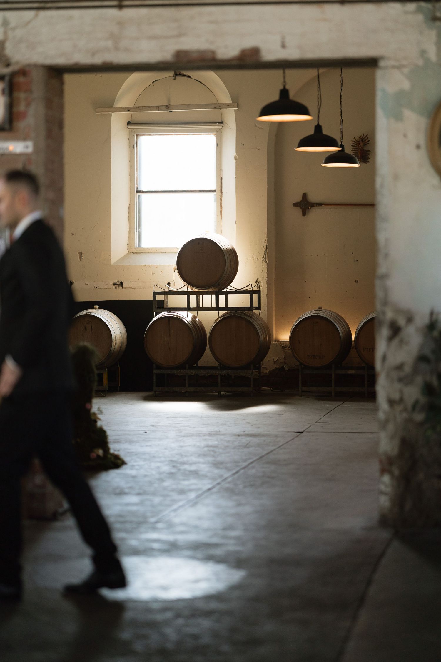 Rustic winery interior with wooden barrels stacked on racks, pendant lights, and a man in a suit walking by.