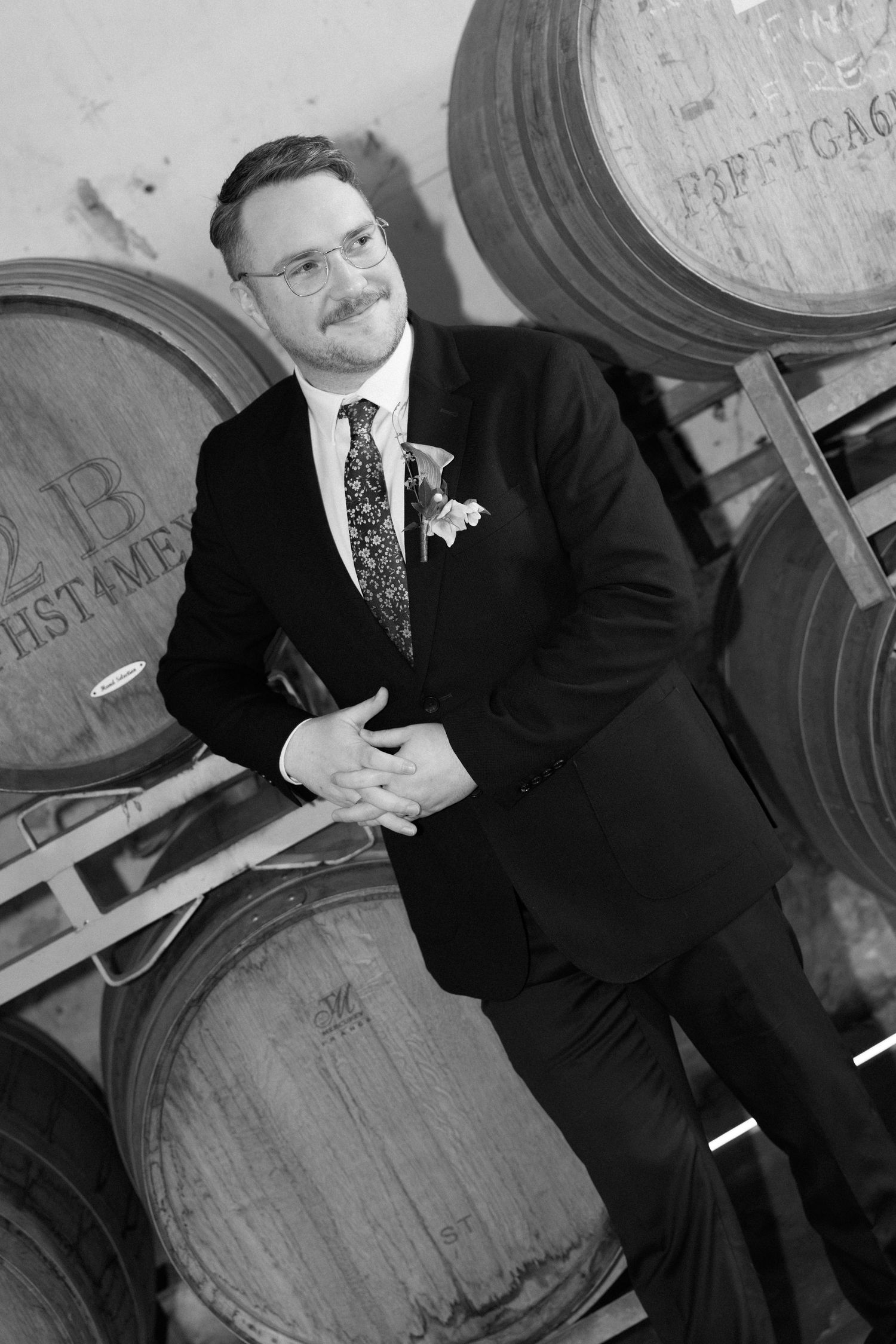 Dapper groom in suit posing beside oak wine barrels in winery in black and white portrait.