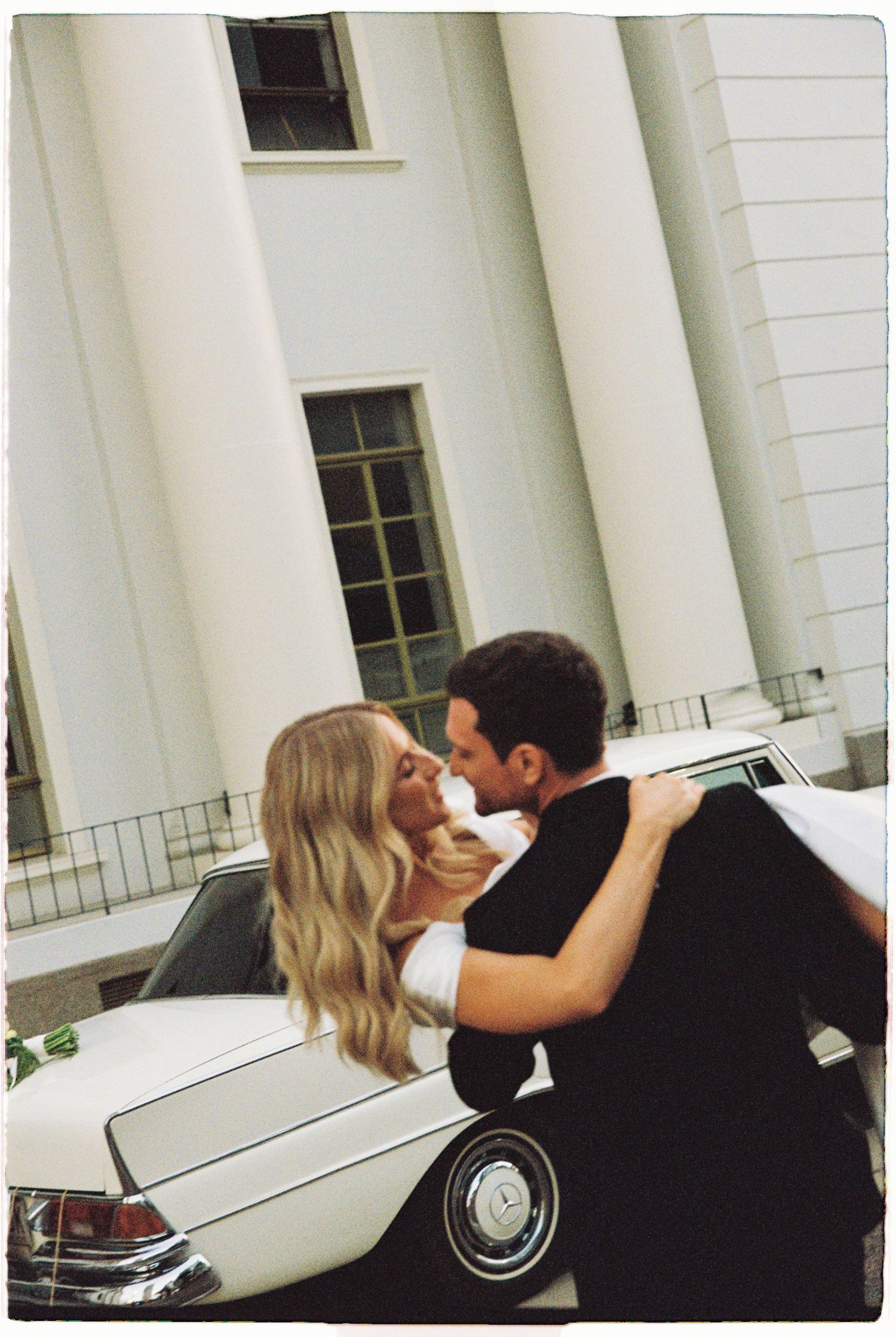 Bride and groom sharing a romantic kiss beside a classic white vintage car at wedding.