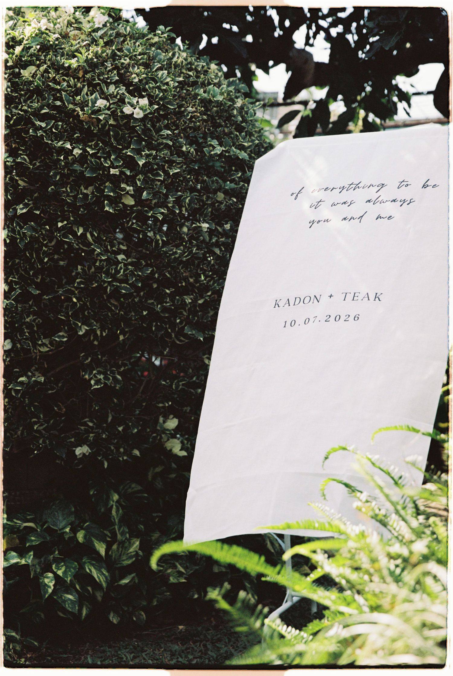 White wedding sign with cursive quote and names Kadon and Teak dated 10-07-2026 leaning against lush green hedge.