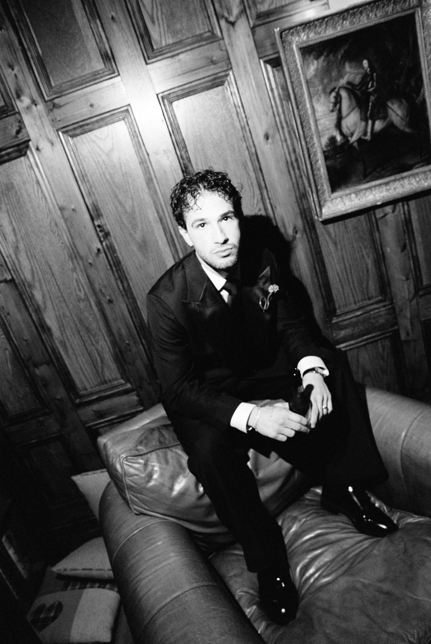 Dapper groom in black suit sitting on leather sofa beside equestrian portrait.
