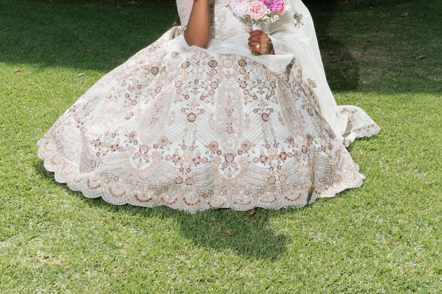 Close-up of bride's stunning white embroidered Indian lehenga skirt with intricate floral lace detailing on grass.
