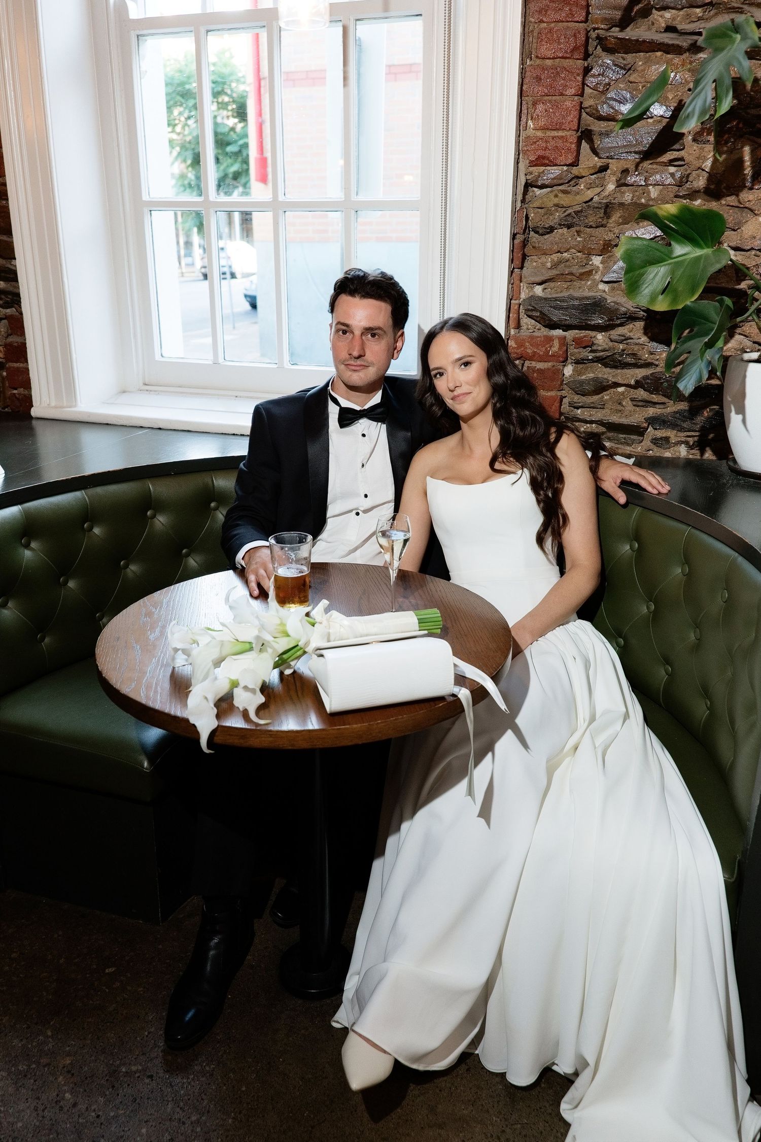 Elegant bride and groom seated at a round table on a green leather booth in a brick-walled bar.