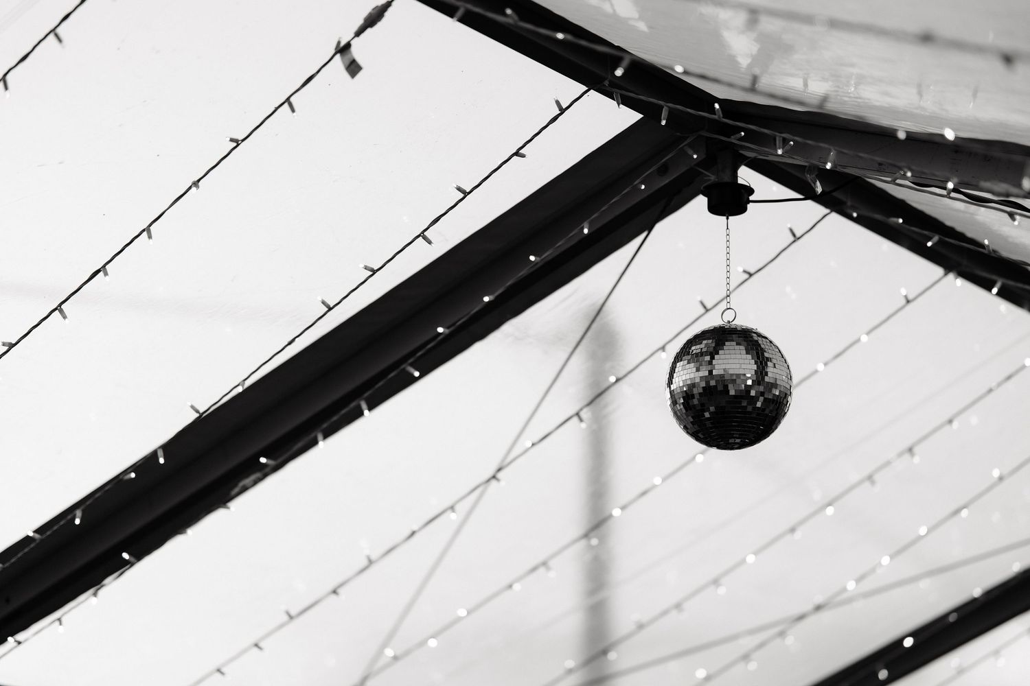 Black and white photo of a disco ball hanging from a ceiling with string lights at a wedding venue.