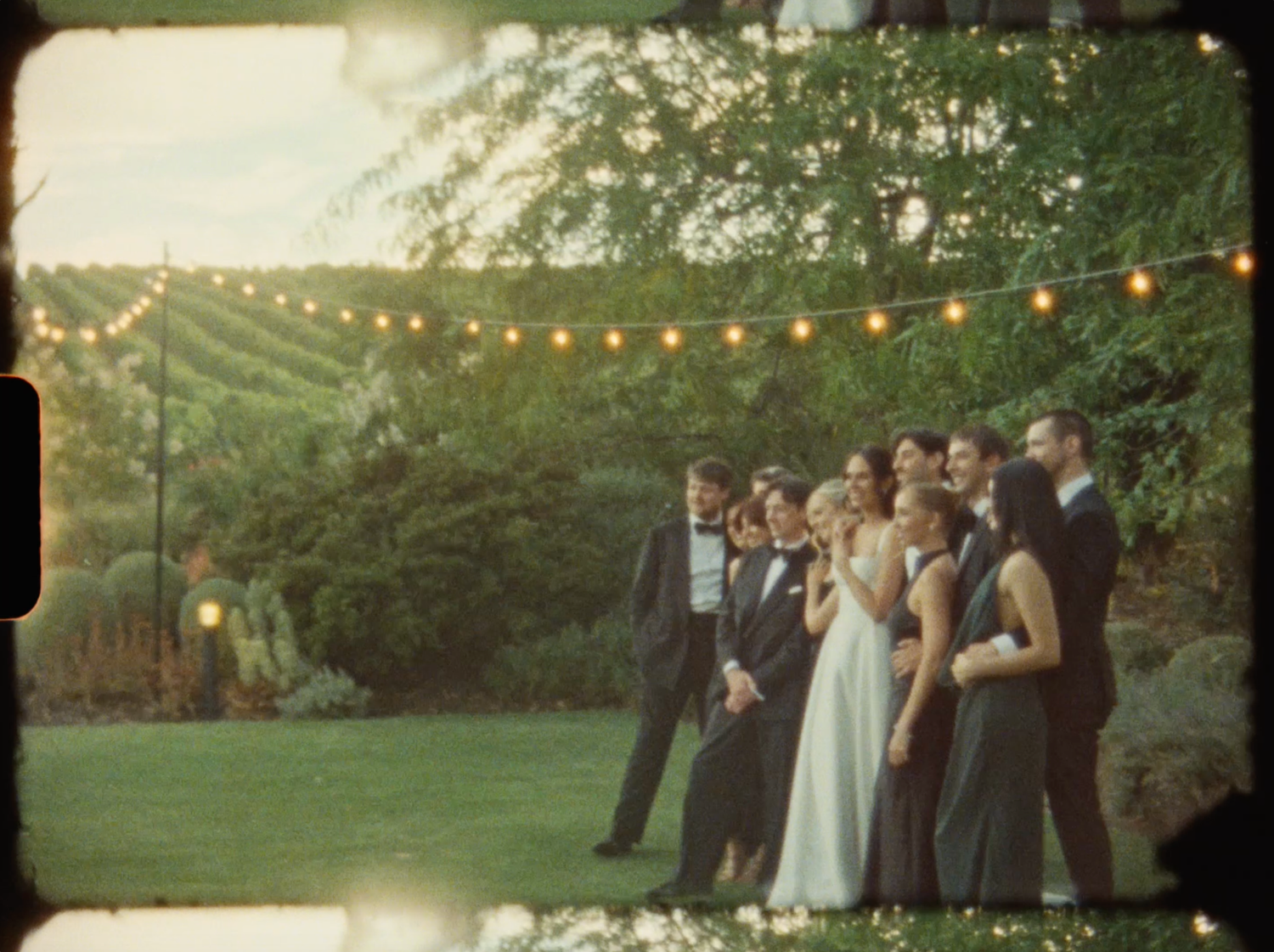 Wedding party poses together under string lights in a lush garden at dusk during outdoor reception.
