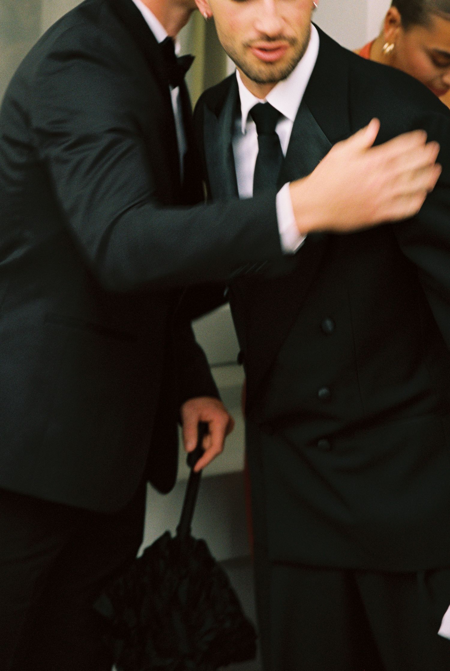 Two men in black suits at formal wedding event, one adjusting the other's jacket lapel.