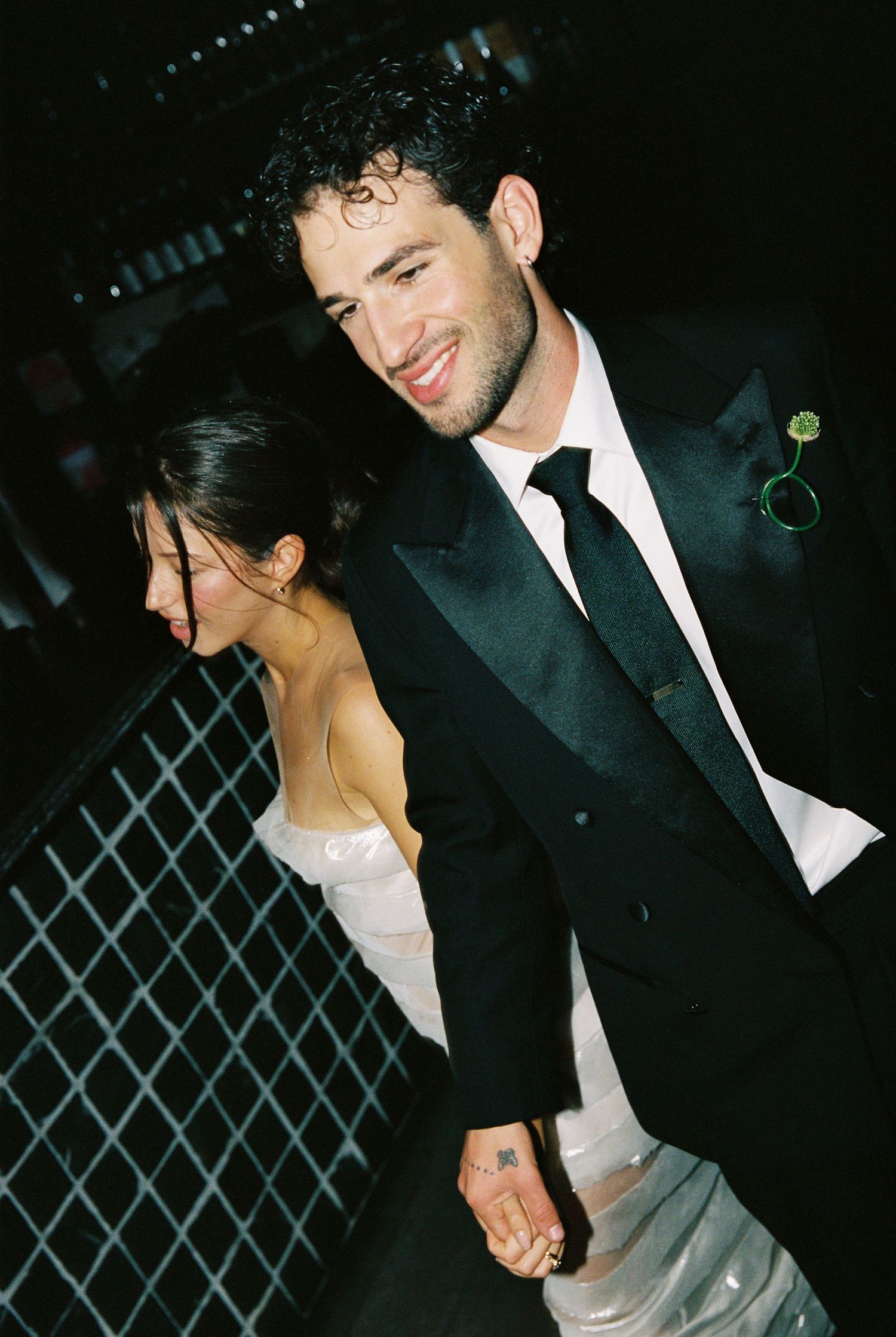 Smiling groom in black tuxedo holds hands with bride in white dress at an evening wedding celebration.