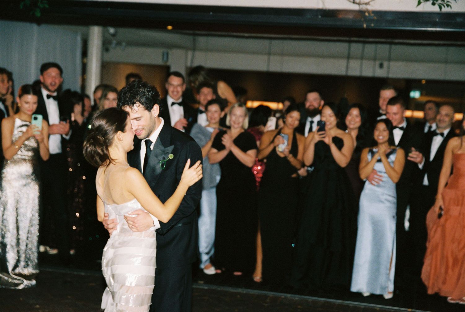 Bride and groom share first dance at elegant wedding reception while guests applaud and photograph the moment.