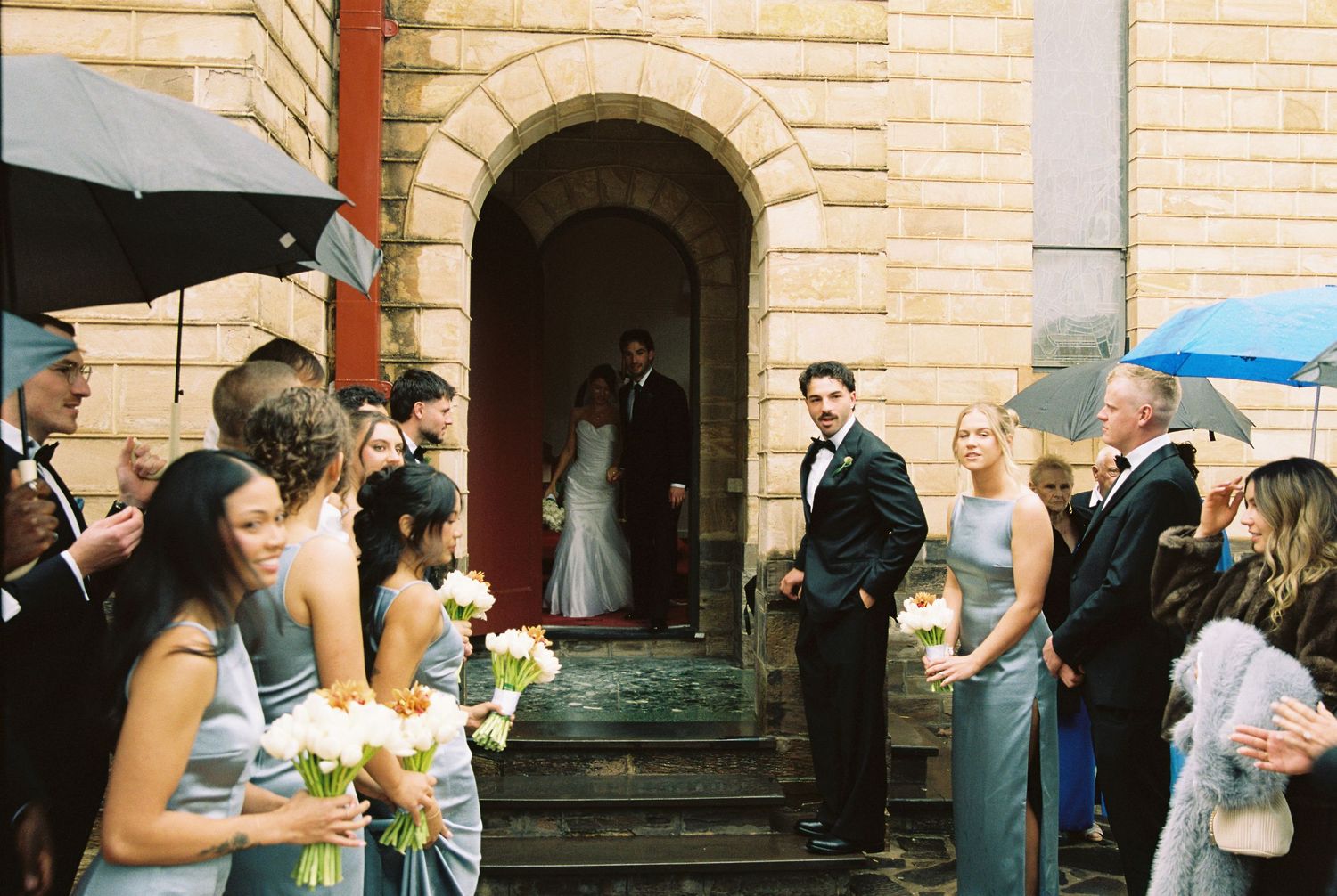 Wedding party exits stone church as bridesmaids in gray dresses hold bouquets while guests gather under umbrellas on rainy day.