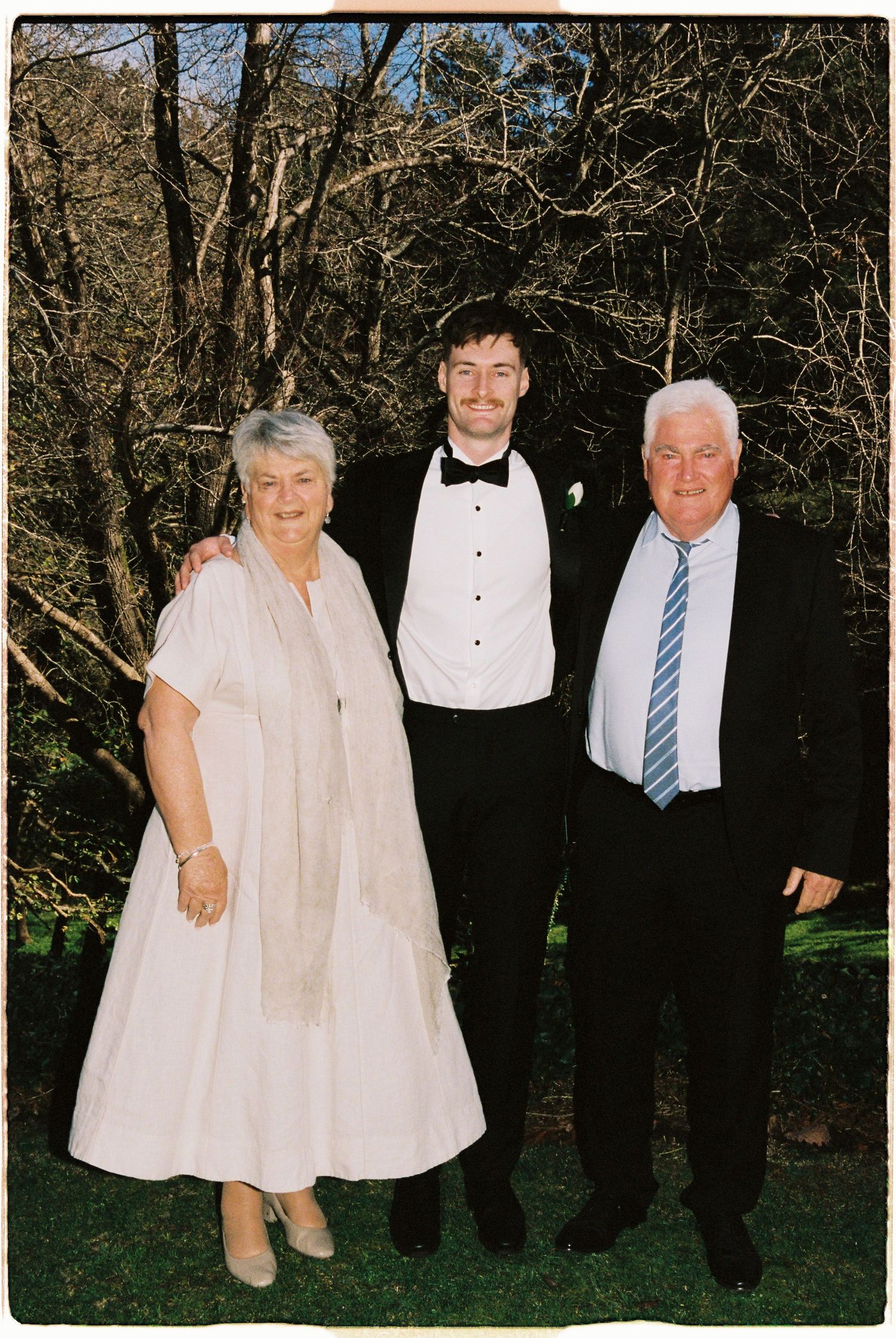 Family of three posing outdoors at a wedding, woman in white dress, young man in tuxedo, older man in suit and tie.