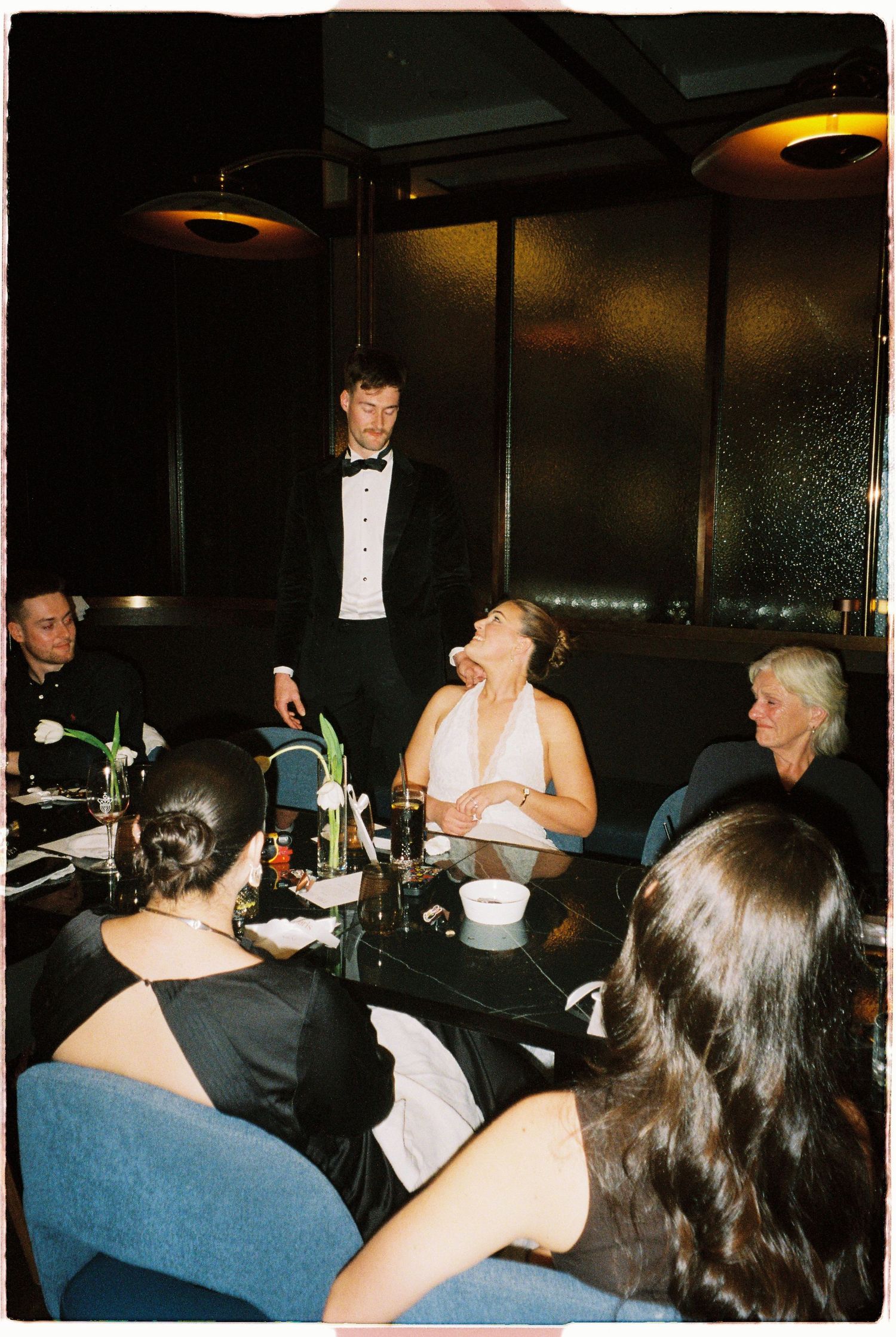 Groom standing at dark elegant reception dinner table while bride laughs joyfully with seated guests.