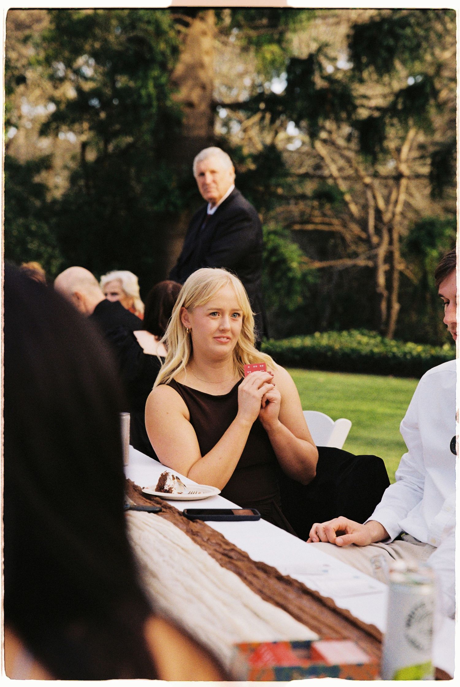 Blonde woman in black dress seated at outdoor wedding reception table in garden.