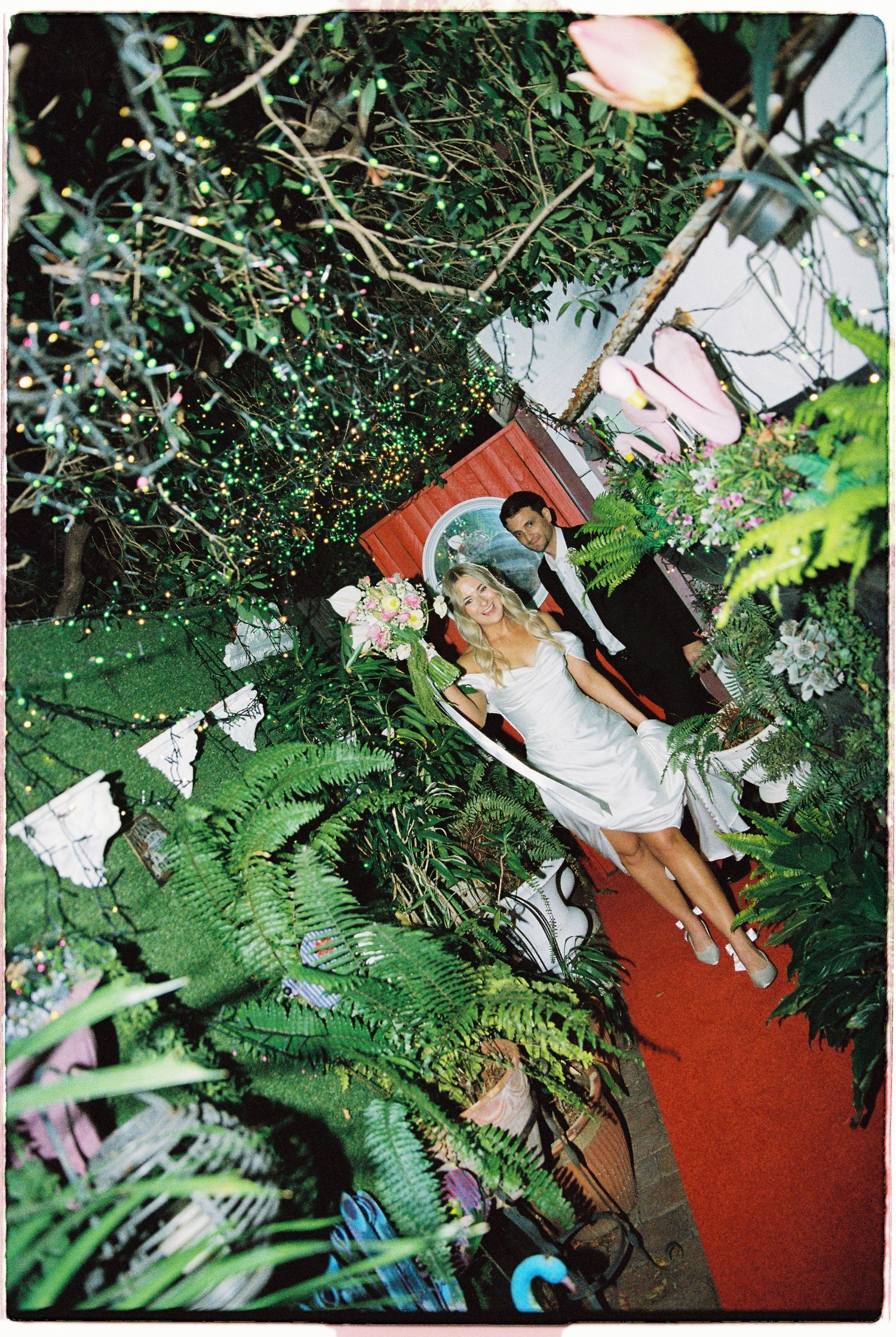 Bride and groom surrounded by lush green ferns and plants at an outdoor garden wedding celebration.
