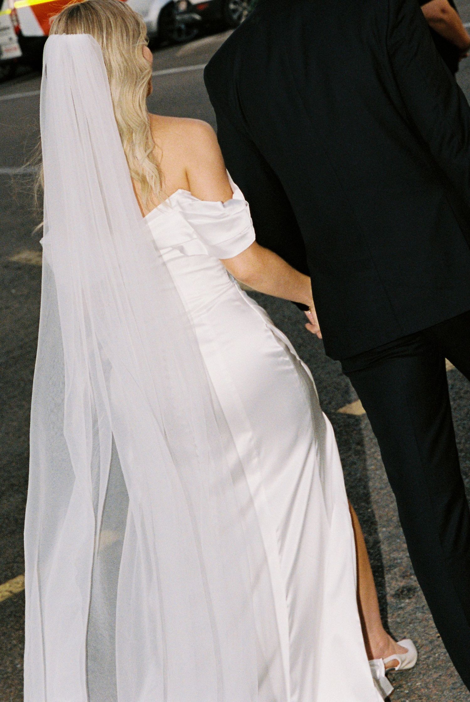 Elegant bride in a fitted white off-shoulder satin gown with long veil walks alongside groom in black suit.
