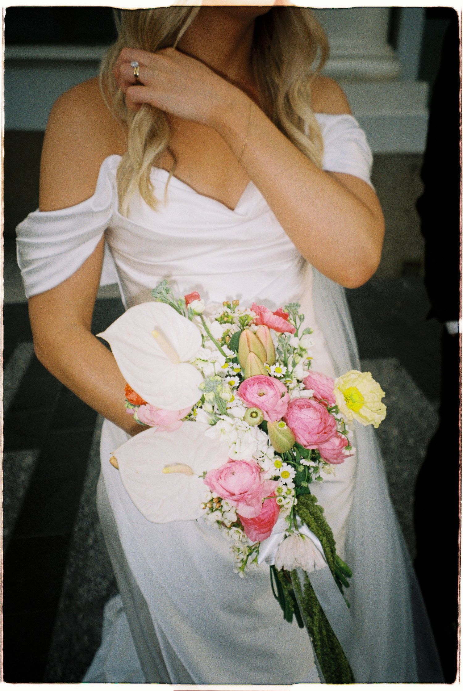 Bride in off-shoulder white gown holding colorful spring bouquet with pink ranunculus and calla lilies.