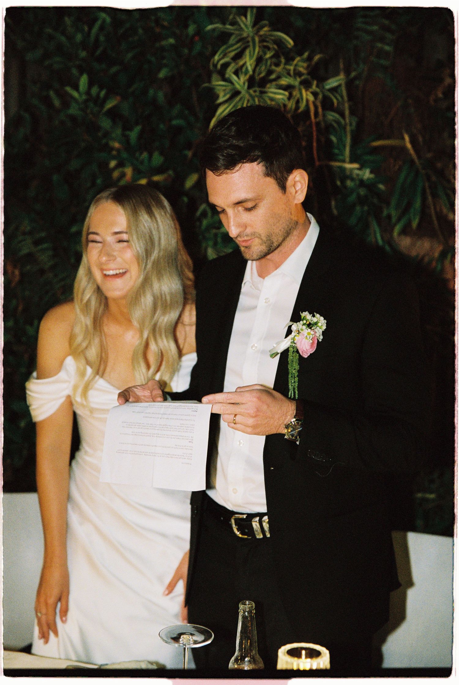 Bride and groom reading wedding vows together, bride in white off-shoulder gown, groom in black suit with floral boutonniere.