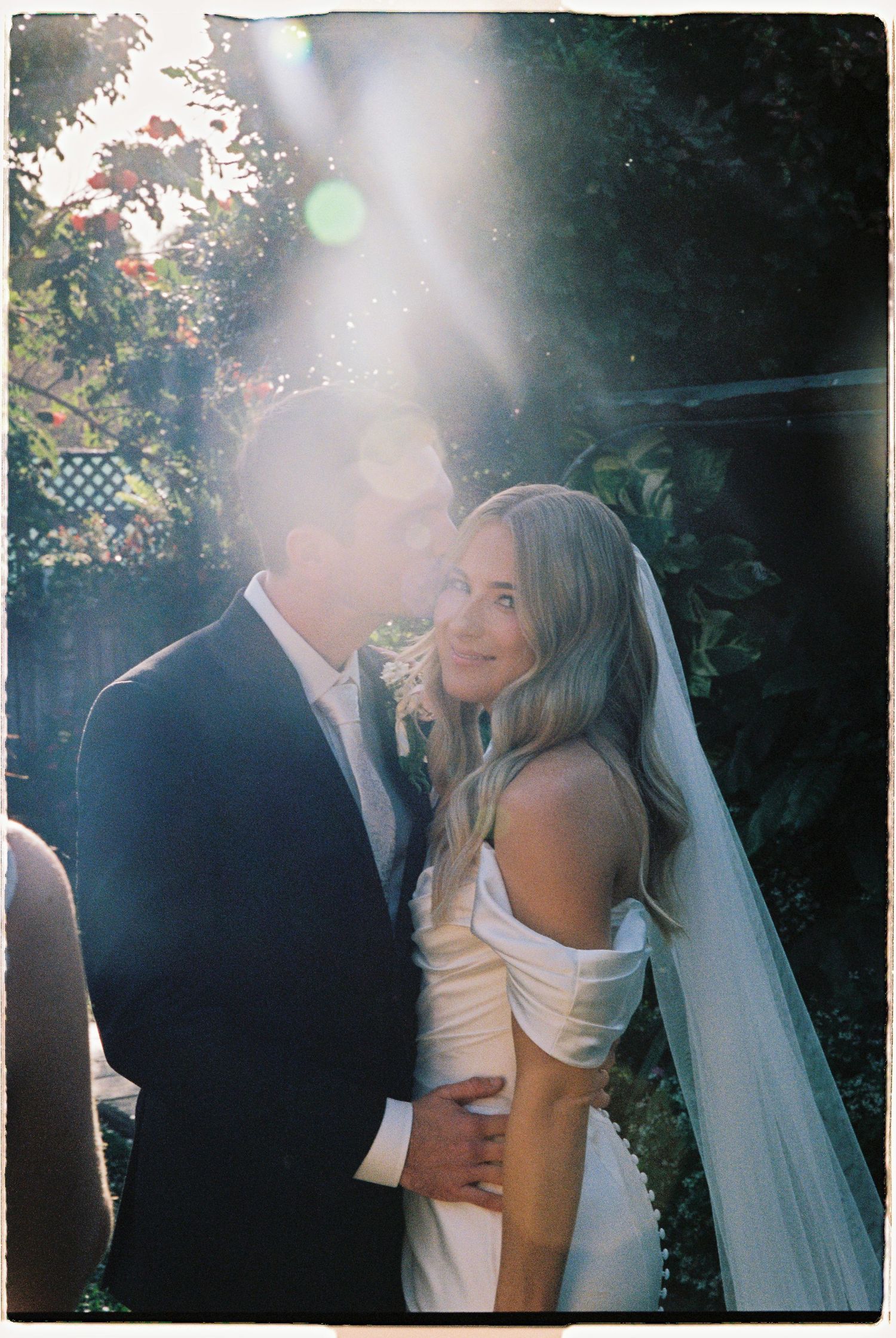 Bride and groom share a romantic embrace in sunlit garden, bride wearing off-shoulder white gown and veil.