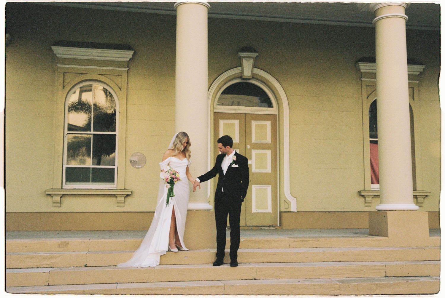 Bride and groom stand on steps of elegant colonial building with white columns, exchanging vows outdoors.