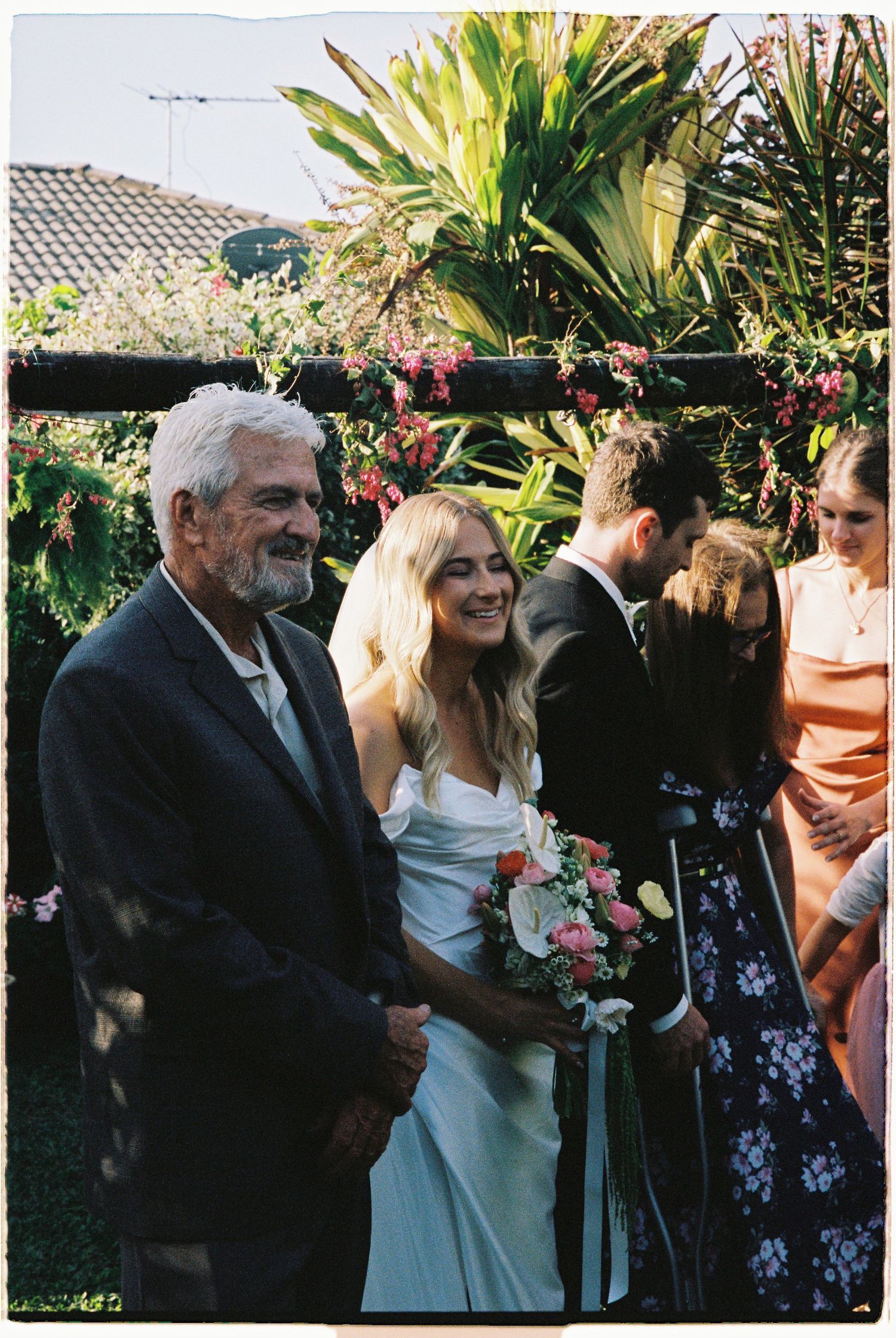 Bride smiling with bouquet walks down aisle with elderly man at outdoor garden wedding ceremony.