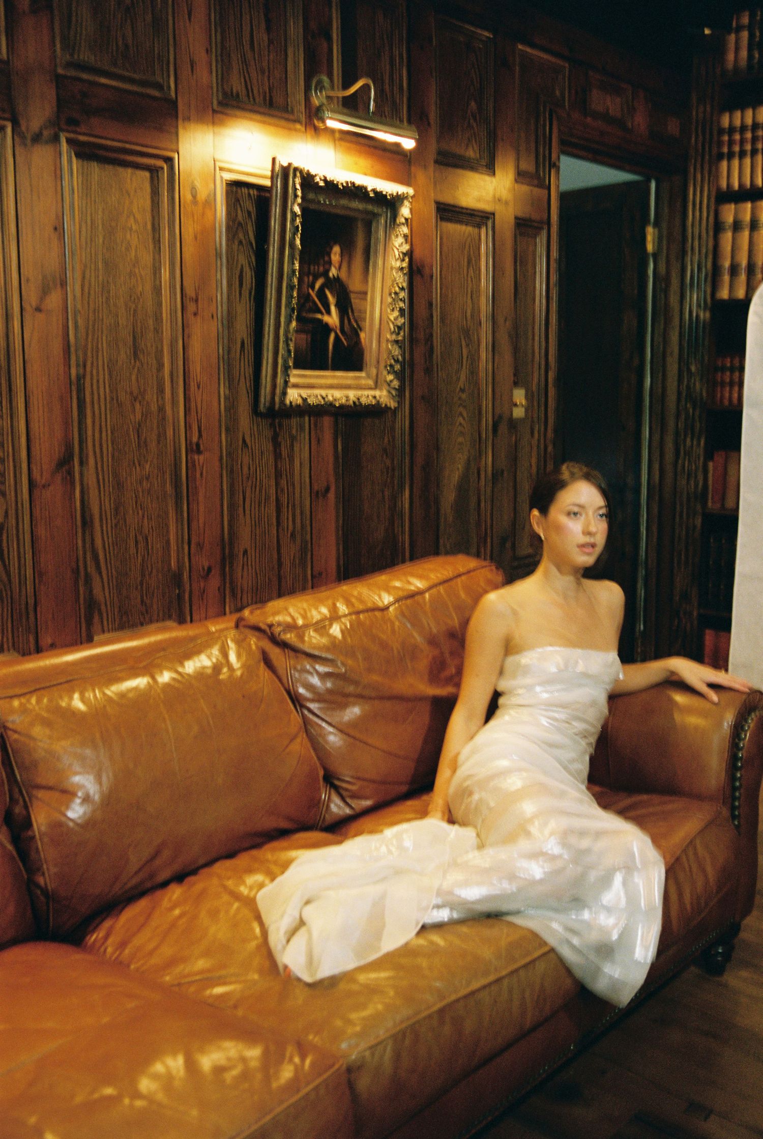 Bride in white strapless gown seated elegantly on a leather sofa in a wood-paneled room.