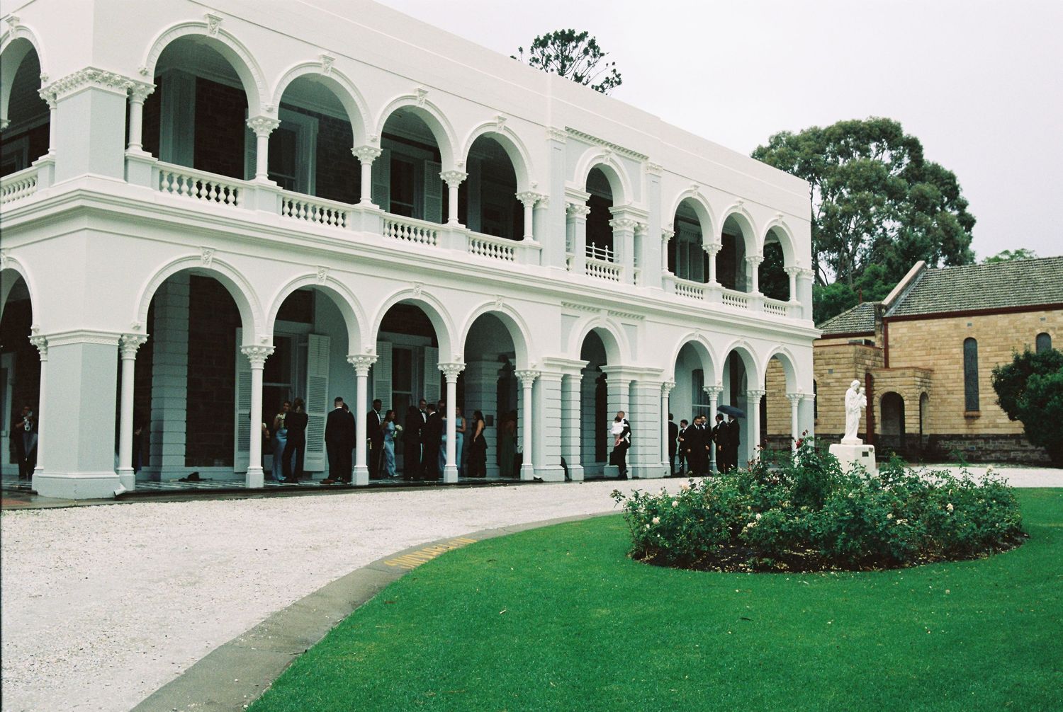 Elegant white colonial building with arched colonnades, lush green lawn, and circular garden bed on a sunny day.