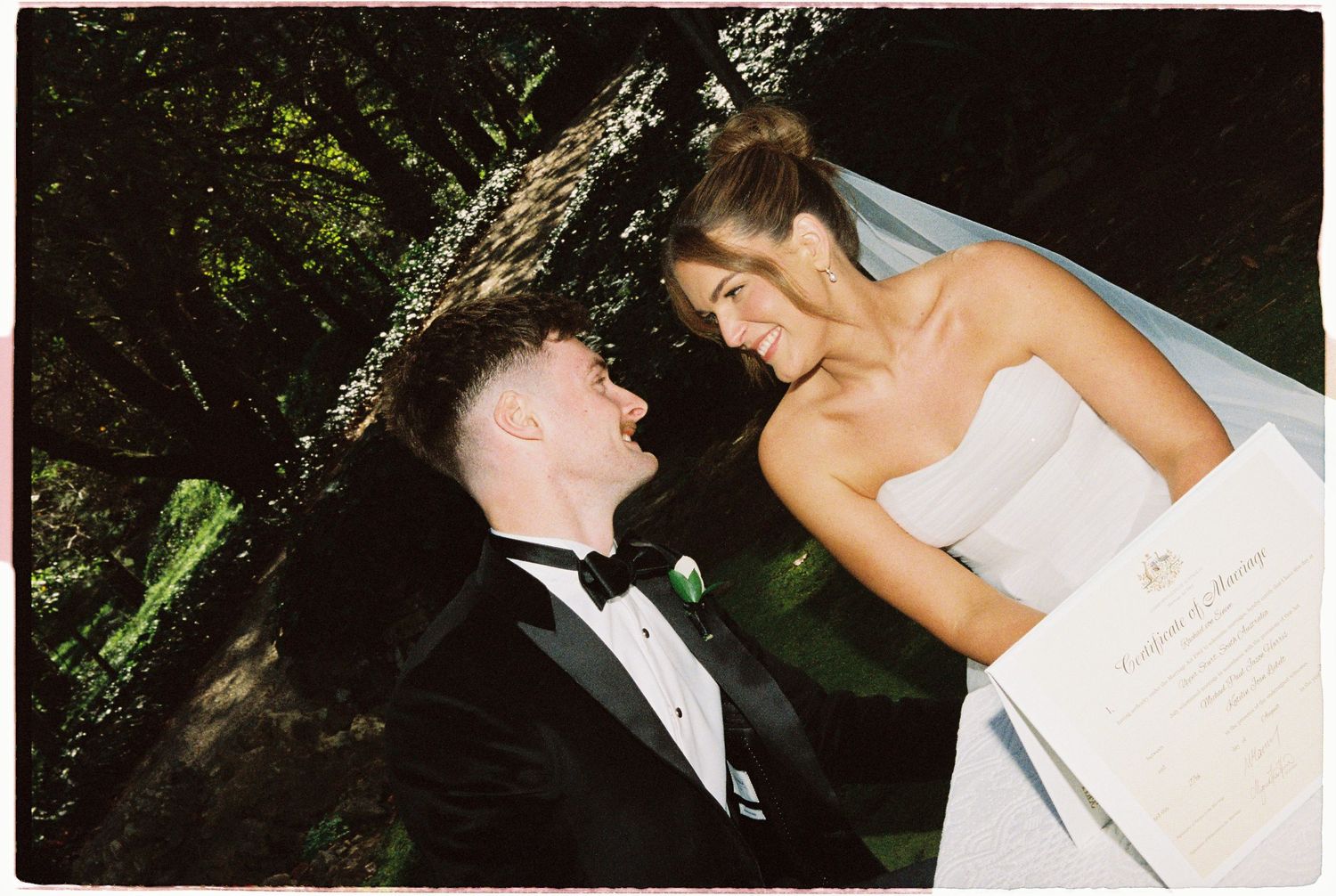 A joyful bride in a strapless gown and groom in a tuxedo share a laughing moment outdoors on their wedding day.