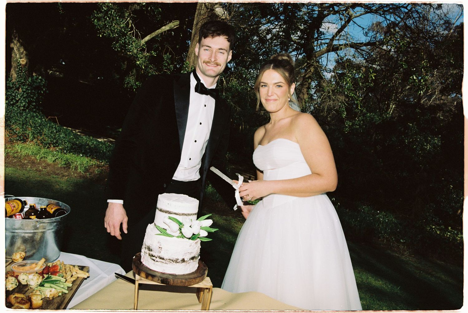 Bride and groom cutting their white wedding cake decorated with green foliage outdoors.