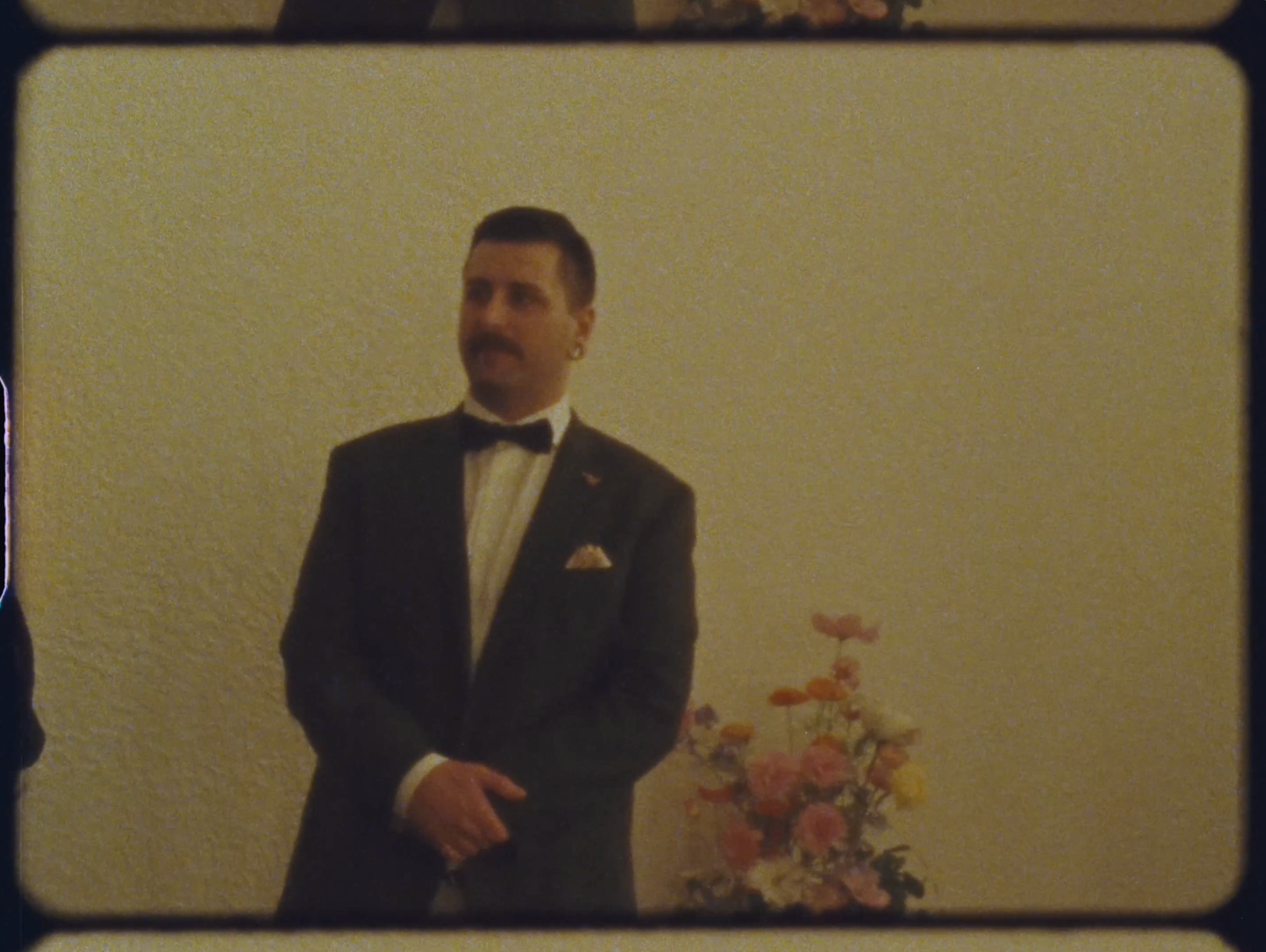 A man in a black tuxedo with mustache stands near a floral arrangement at a formal event.