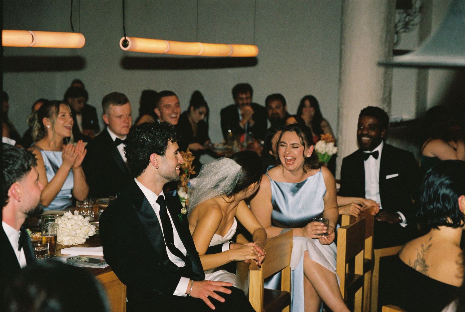 Wedding guests laugh and celebrate at reception dinner table, with bridesmaids in blue dresses and groomsmen in tuxedos.