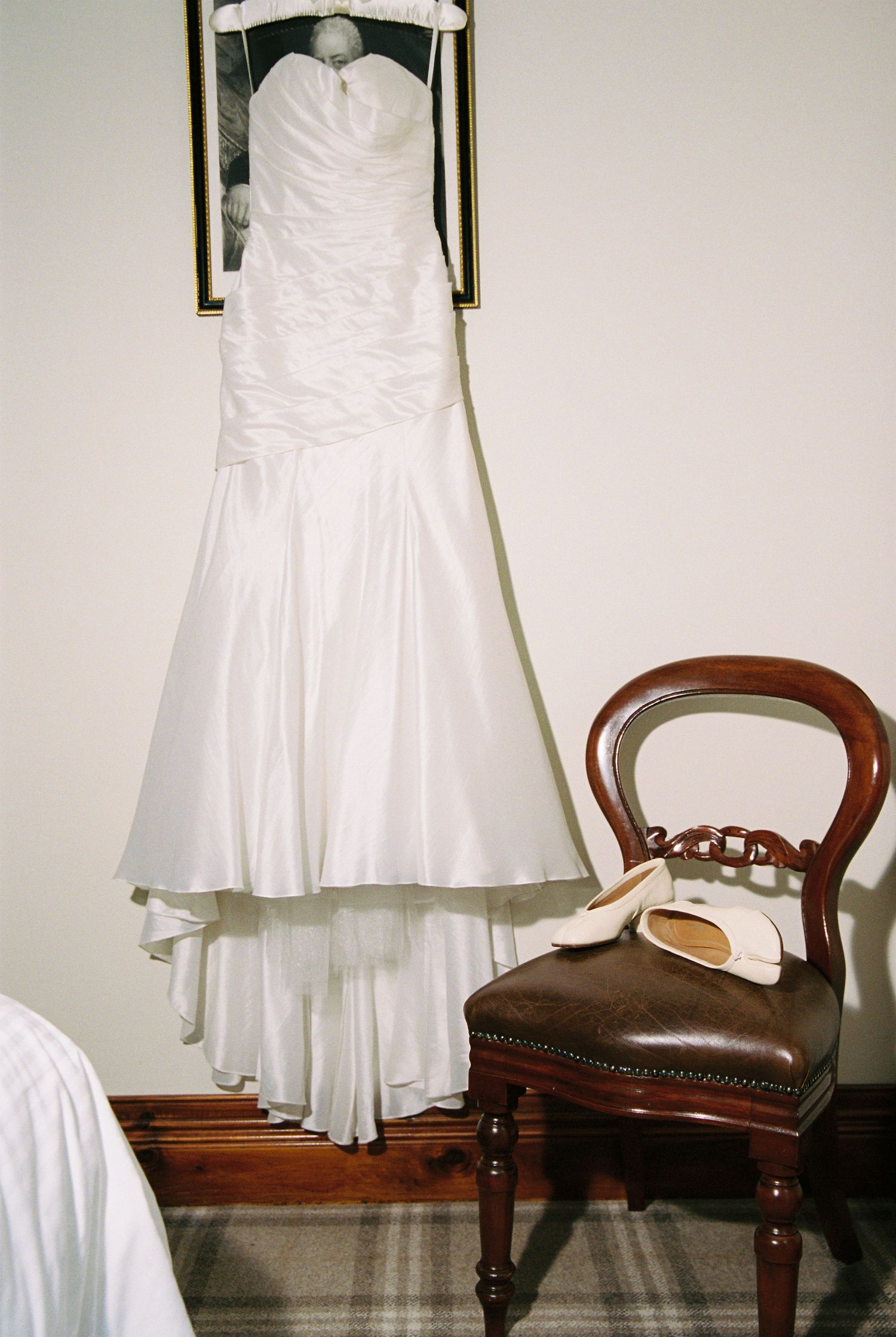 Elegant strapless white wedding gown hanging on wall above vintage wooden chair with bridal shoes.