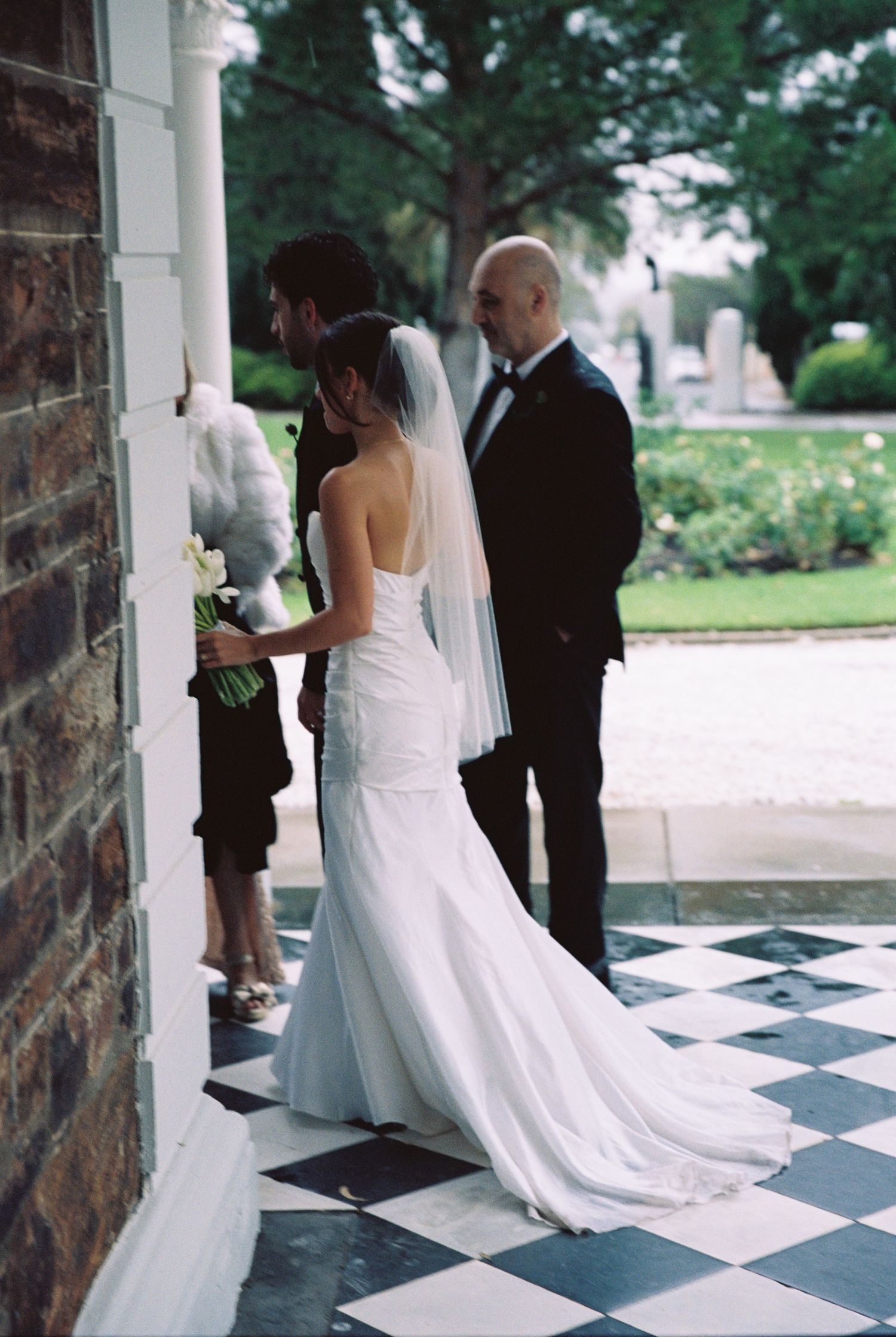 Bride in white gown and veil stands with bald groom on checkered floor at outdoor wedding venue.